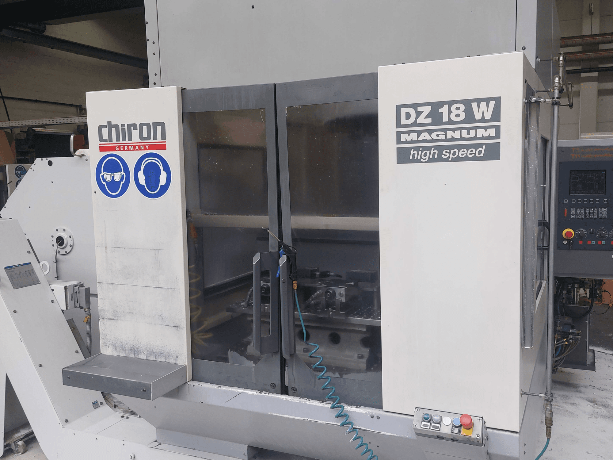 Vertikalus apdirbimo centras „CHIRON DZ 18 W MAGNUM“, vaizdas iš priekio, kuriame matomas valdymo pultas ir saugos ženklai.