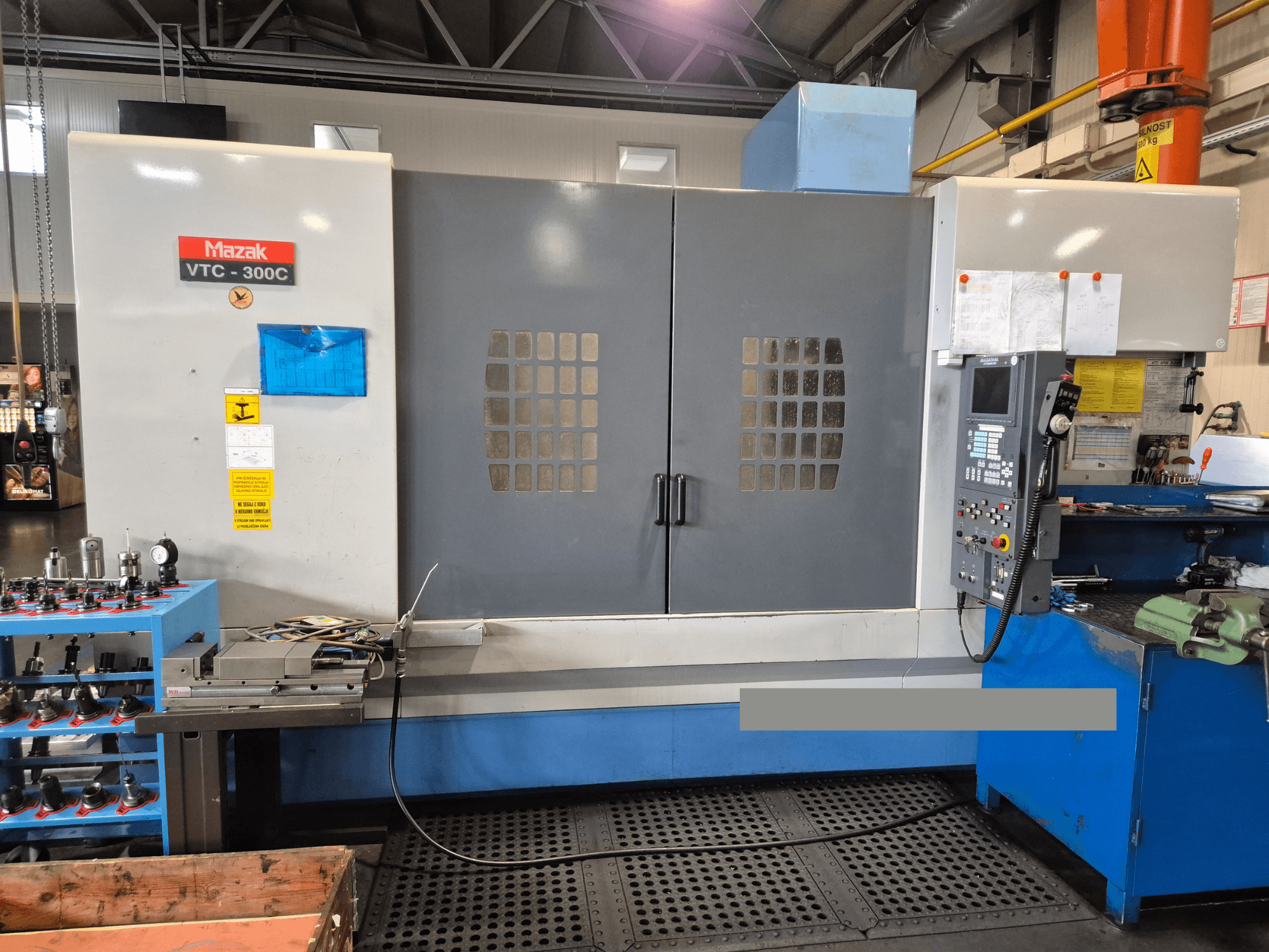 Mazak VTC-300C staklės gamykloje, priekinis vaizdas, kuriame matyti valdymo skydelis, pilkos durys su langų tinkleliu ir šalia esanti įranga.