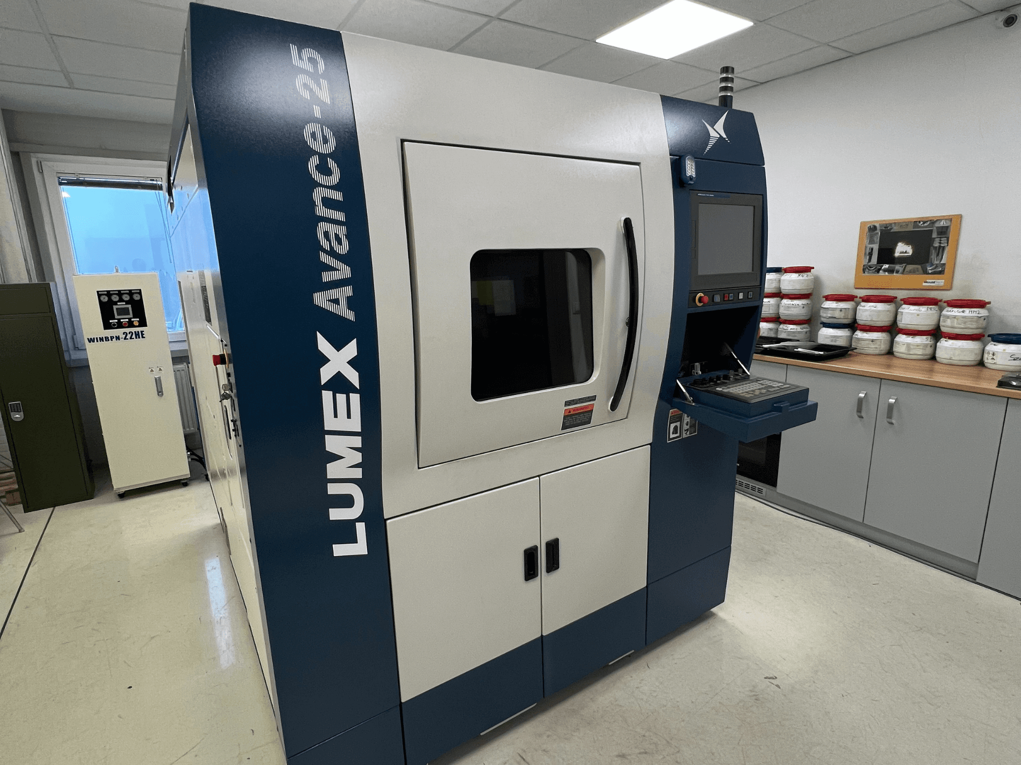 LUMEX Avance-25 3D spausdintuvas laboratorijoje, vaizdas iš priekio. Valdymo skydelis su matomu ekranu ir mygtukais, įranga fone.