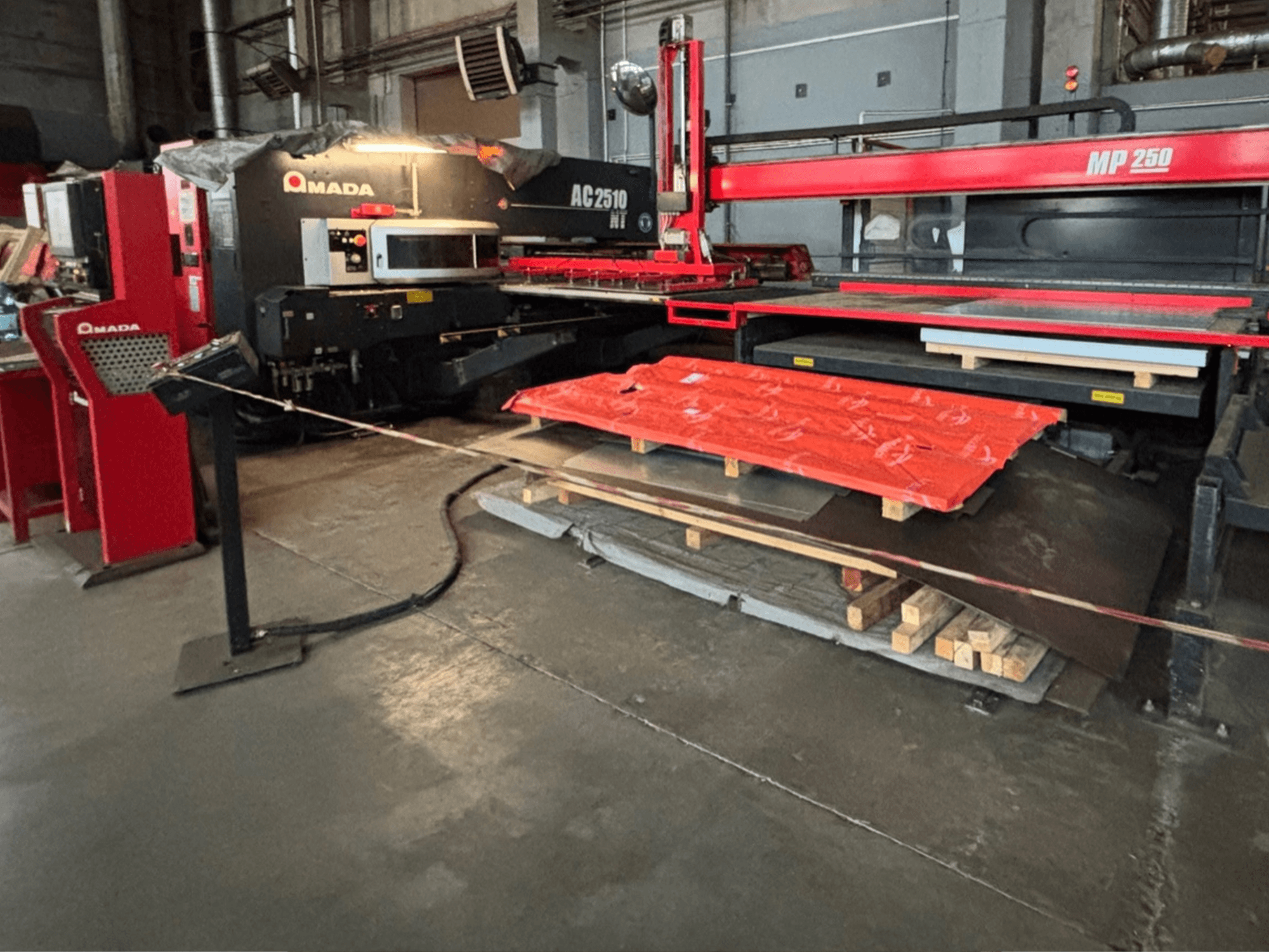 "Amada AC2510 NT" lazerinis pjaustytuvas gamykloje, vaizdas iš priekio, sunkios mašinos su metalo lakštais ir padėklais ant grindų.