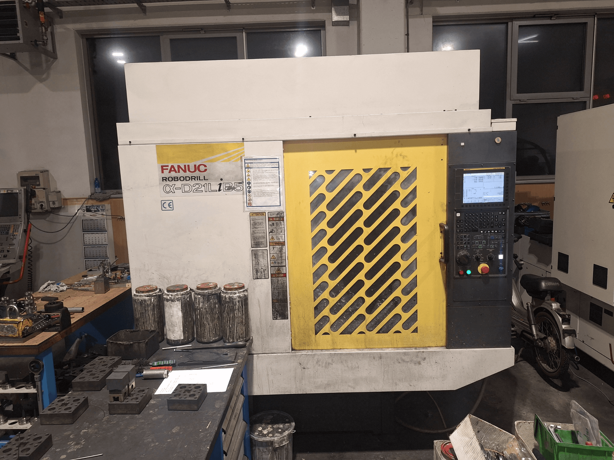 FANUC Robodrill α-D21LiB5 priekinis vaizdas, kuriame matyti valdymo skydelis, geltonos grotelės ir šalia esantys įrankiai ant darbo stalo.