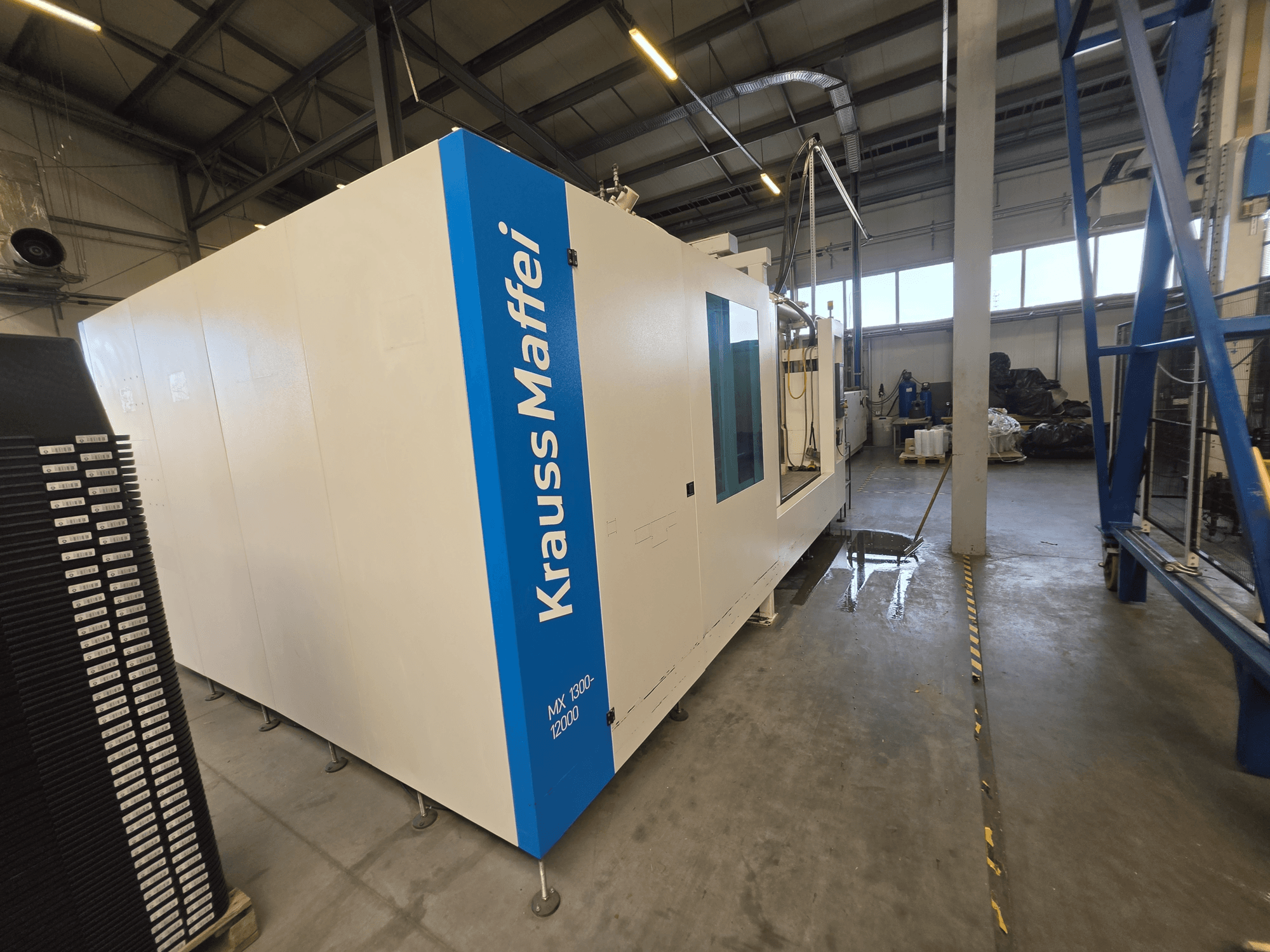 „Krauss Maffei MX 1300-12000“ hidraulinio liejimo staklės, matomos iš šono, su dideliu baltu rėmu ir mėlynu logotipu.