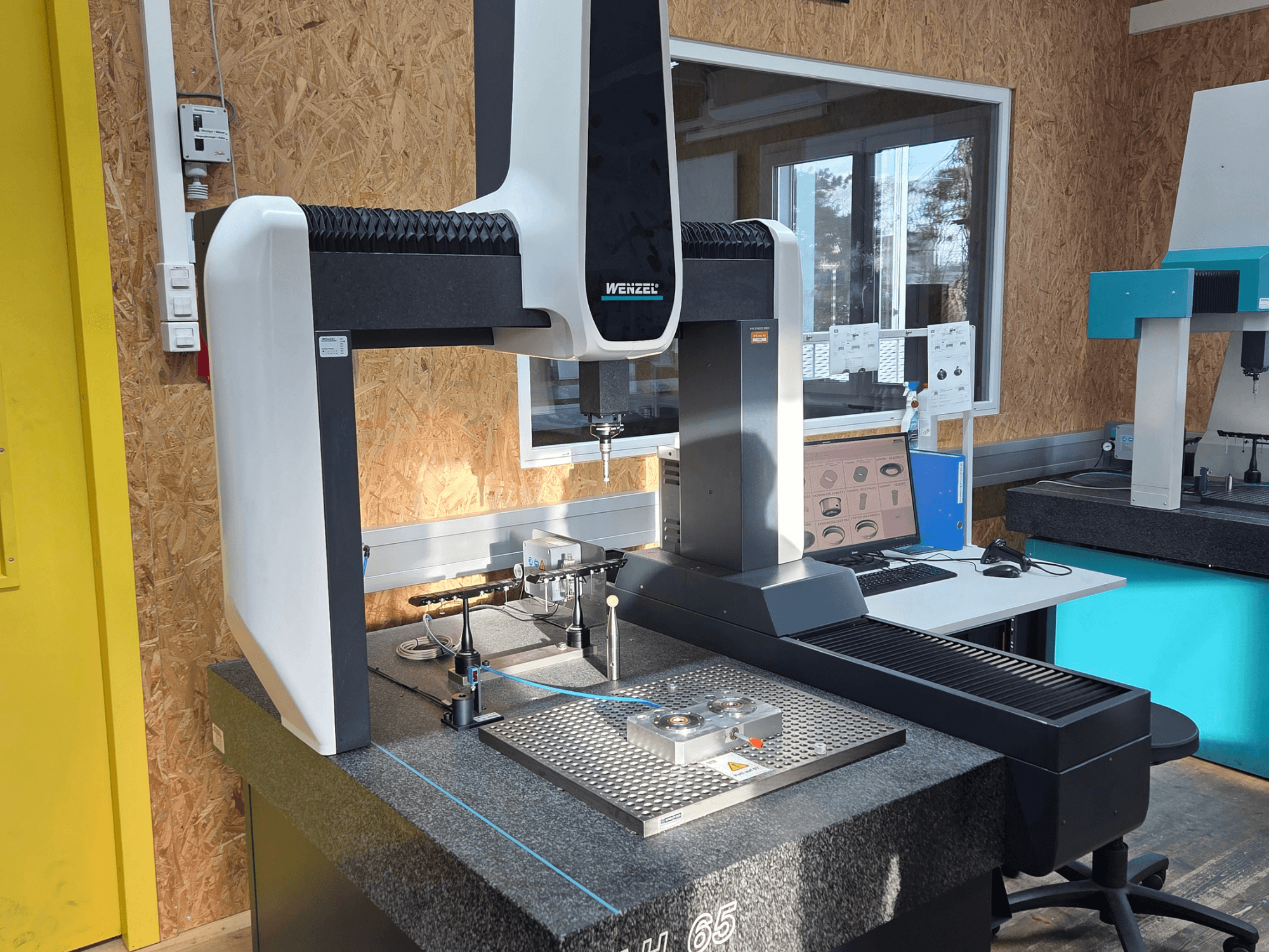 Wenzel LH 65 koordinačių matavimo mašina laboratorijoje, vaizdas iš priekio, matomi matavimo įrankiai ir monitorius.