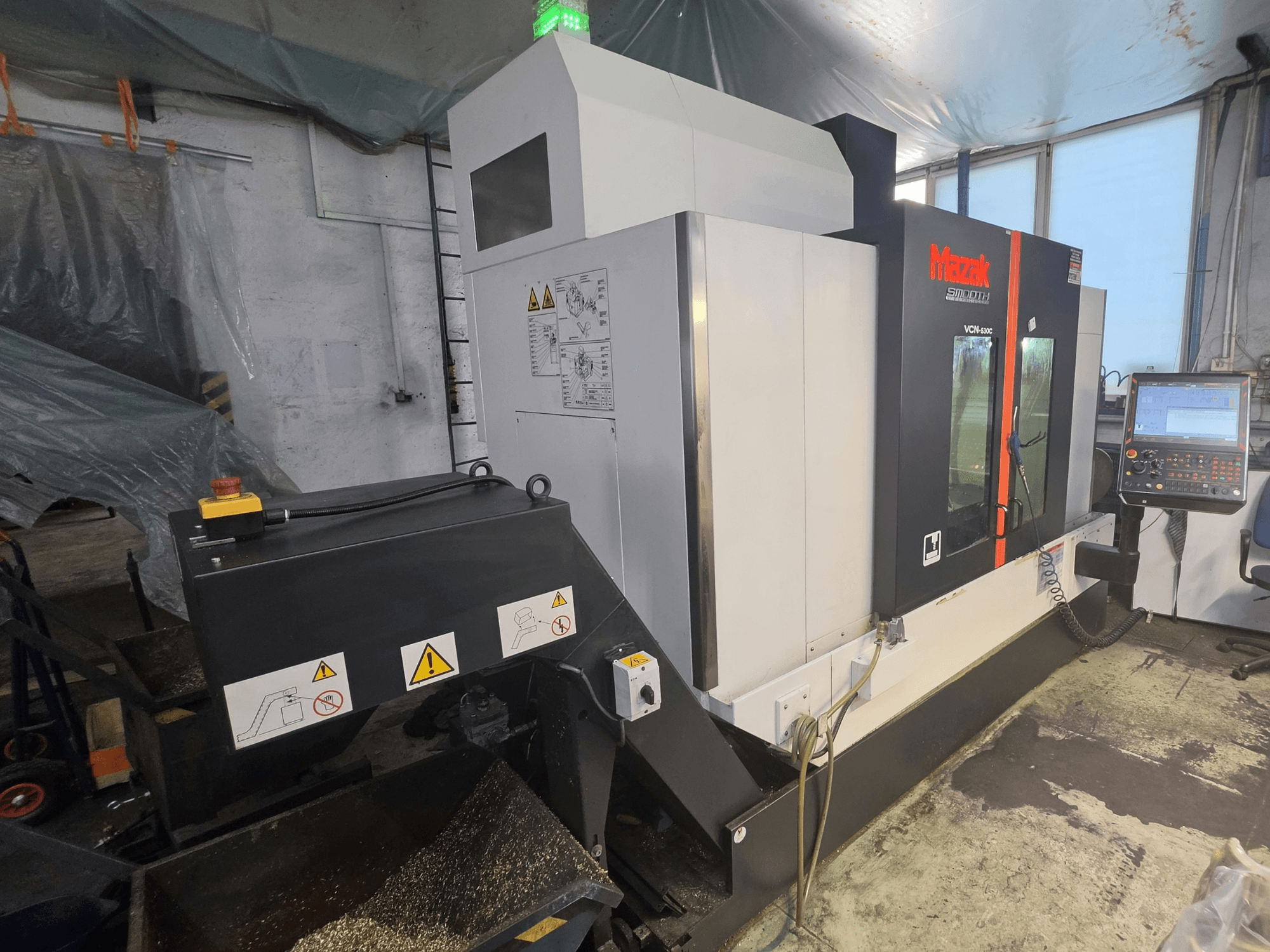 „Mazak VCN-530C“ vertikaliojo apdirbimo centro vaizdas iš priekio, kuriame matomas valdymo skydas, įrankių grandinė ir veikimo indikatoriai.