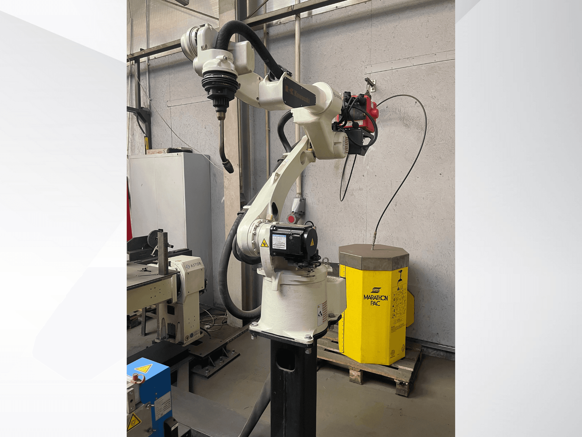 "Kawasaki" roboto rankos šoninis vaizdas su pritvirtintu įrankių laikikliu, fone - geltonas maitinimo blokas.