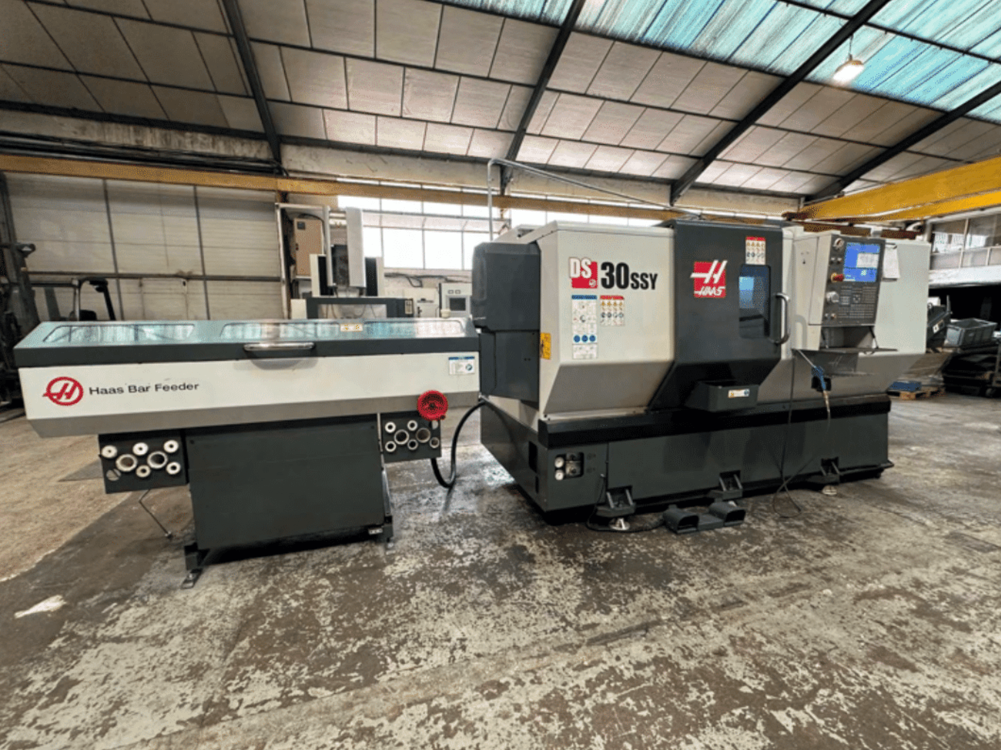 Haas DS-30SSY CNC tekinimo staklės su Haas strypų tiektuvu, šoninis vaizdas, kuriame matomas valdymo skydelis ir tiekimo mechanizmas dirbtuvėse.