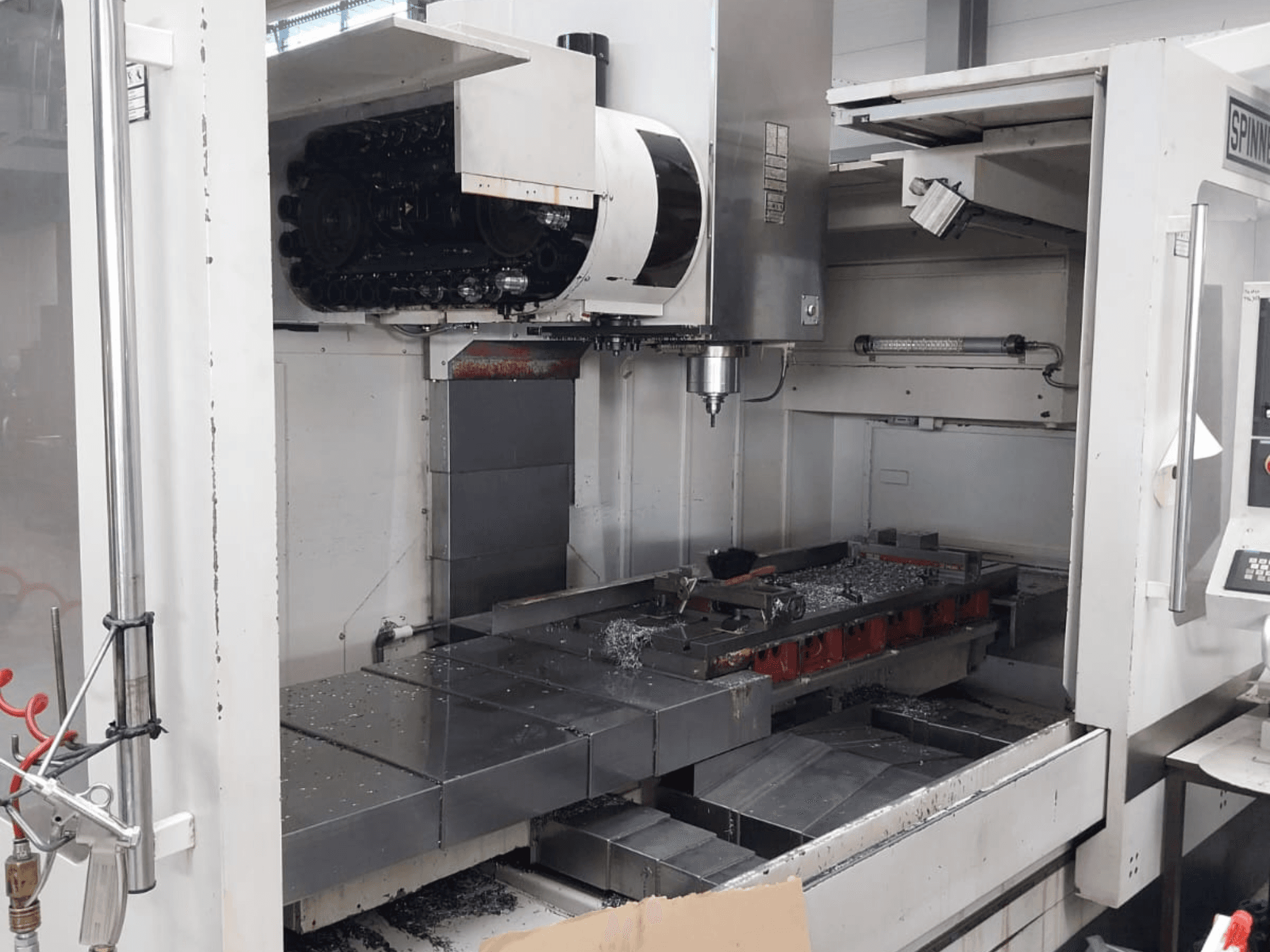 Vertikalaus apdirbimo centro SPINNER MVC 1600 vaizdas iš priekio, kuriame matoma atvira darbo erdvė, suklys ir metalo drožlės ant stalo.