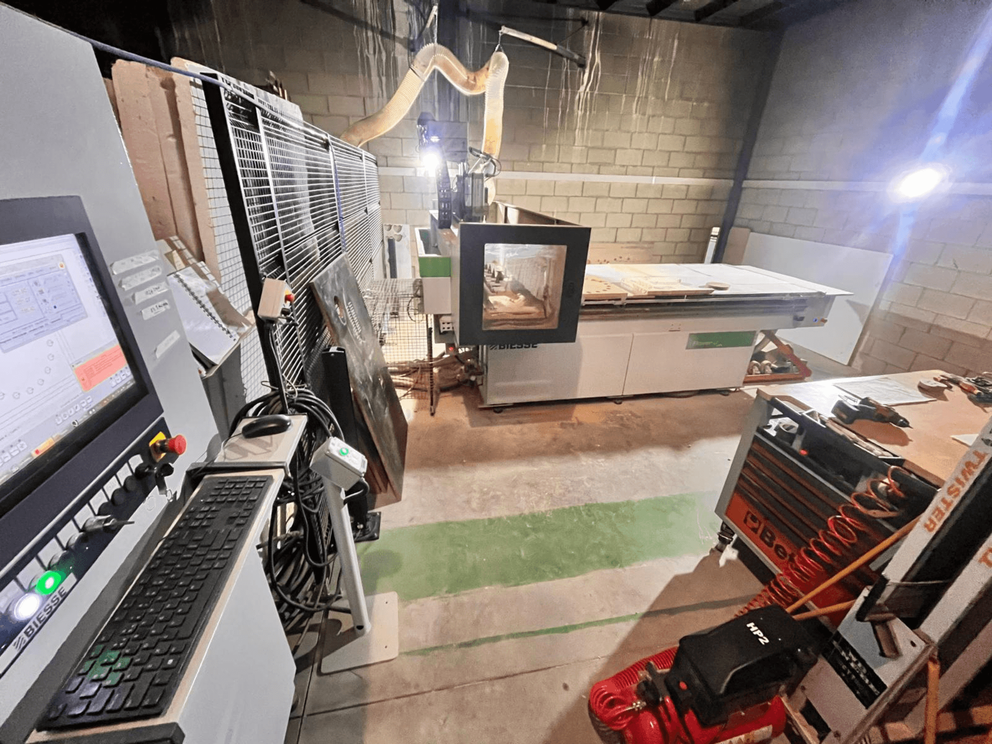 "Biesse" CNC staklės dirbtuvėse, šoninis vaizdas, kuriame matyti valdymo skydelis, dulkių ištraukimas, įrankiai ir įvairios medžiagos ant stalo.