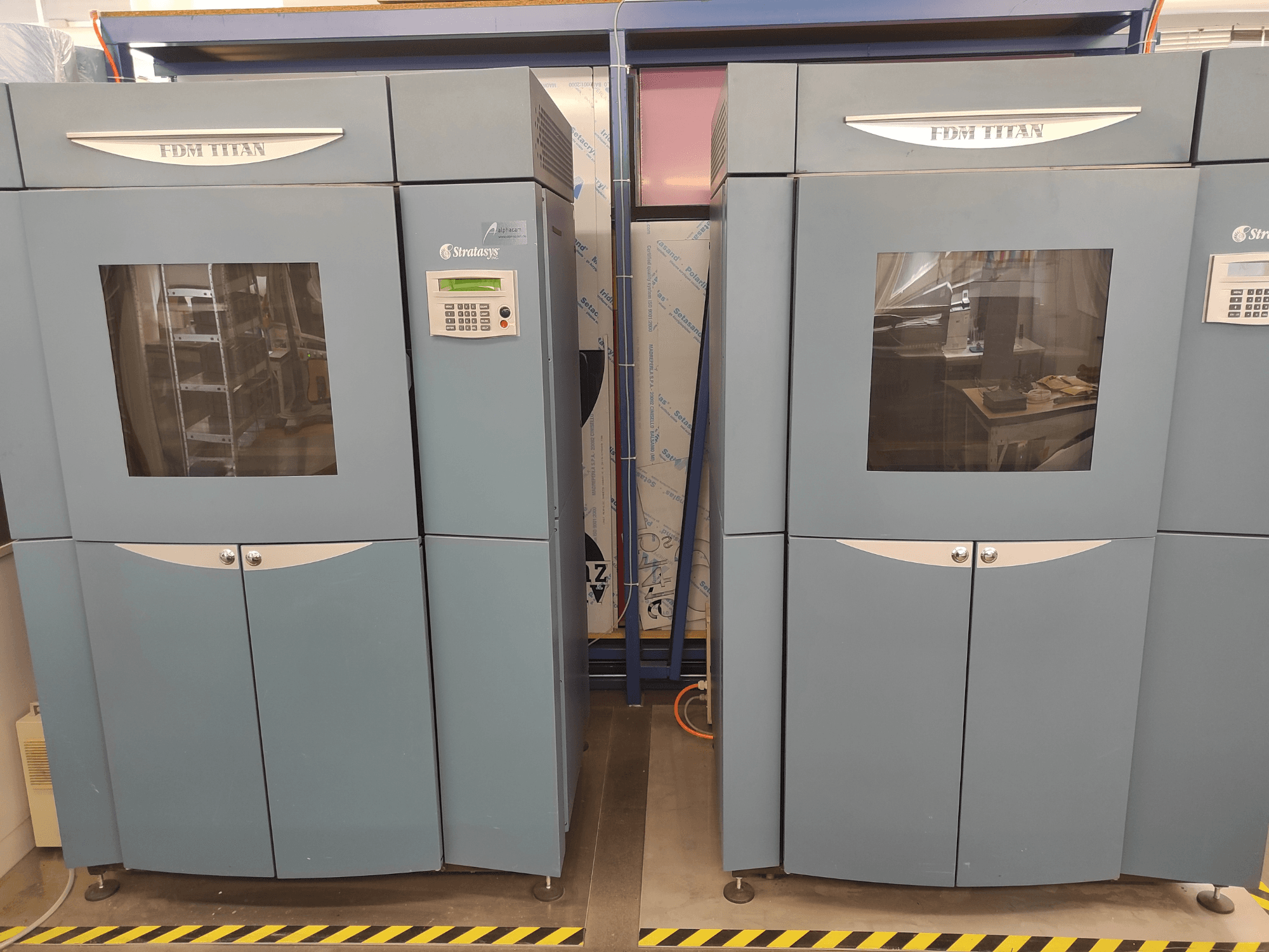 „Stratasys“ gaminamas „FDM TitanTI“ 3D spausdintuvas, vaizdas iš priekio, su dvejomis durelėmis ir valdymo skydeliu, pastatytas pramoninėje aplinkoje.