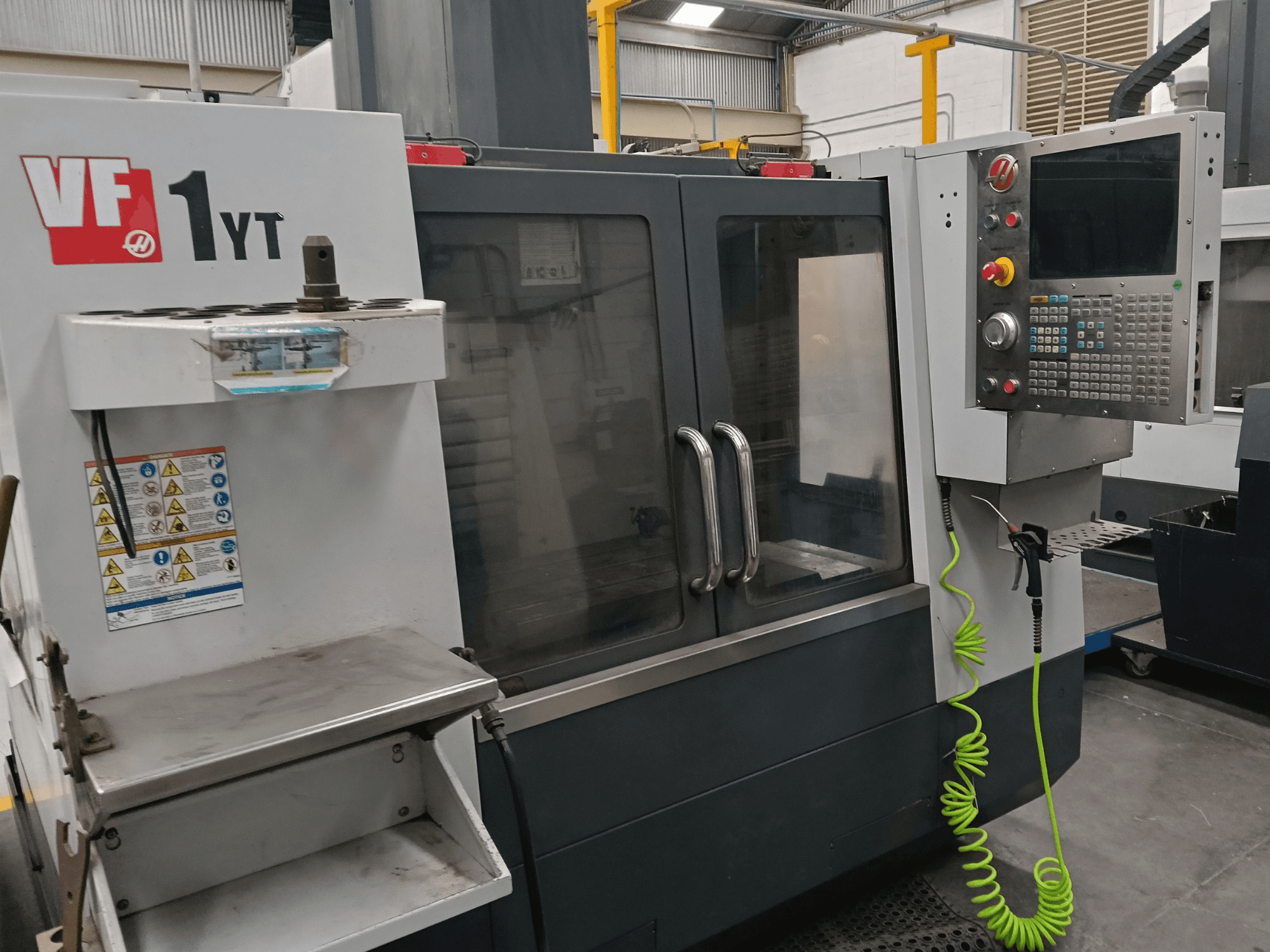 Vertikalusis apdirbimo centras „HAAS VF-1YT-2“, vaizdas iš priekio, kuriame matomas valdymo pultas, įrankių laikiklis, saugos etiketės ir apdirbimo zona.