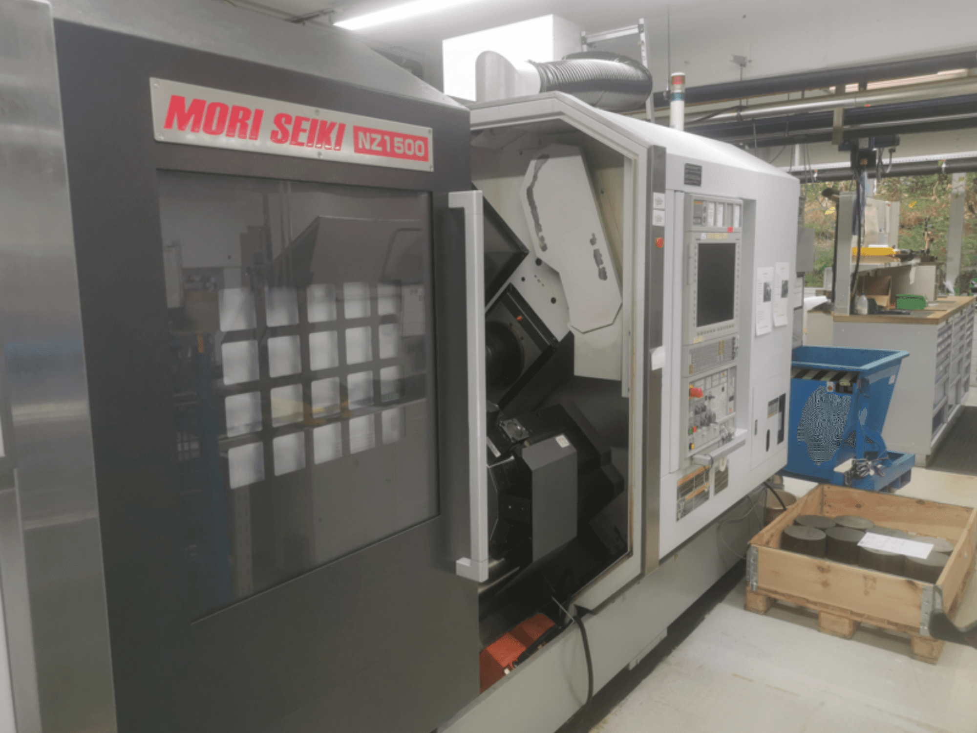 MORI SEIKI NZ1500 CNC staklės nedideliu kampu, matomas valdymo skydelis ir darbo vieta su metaliniais ruošiniais ant medinio padėklo.