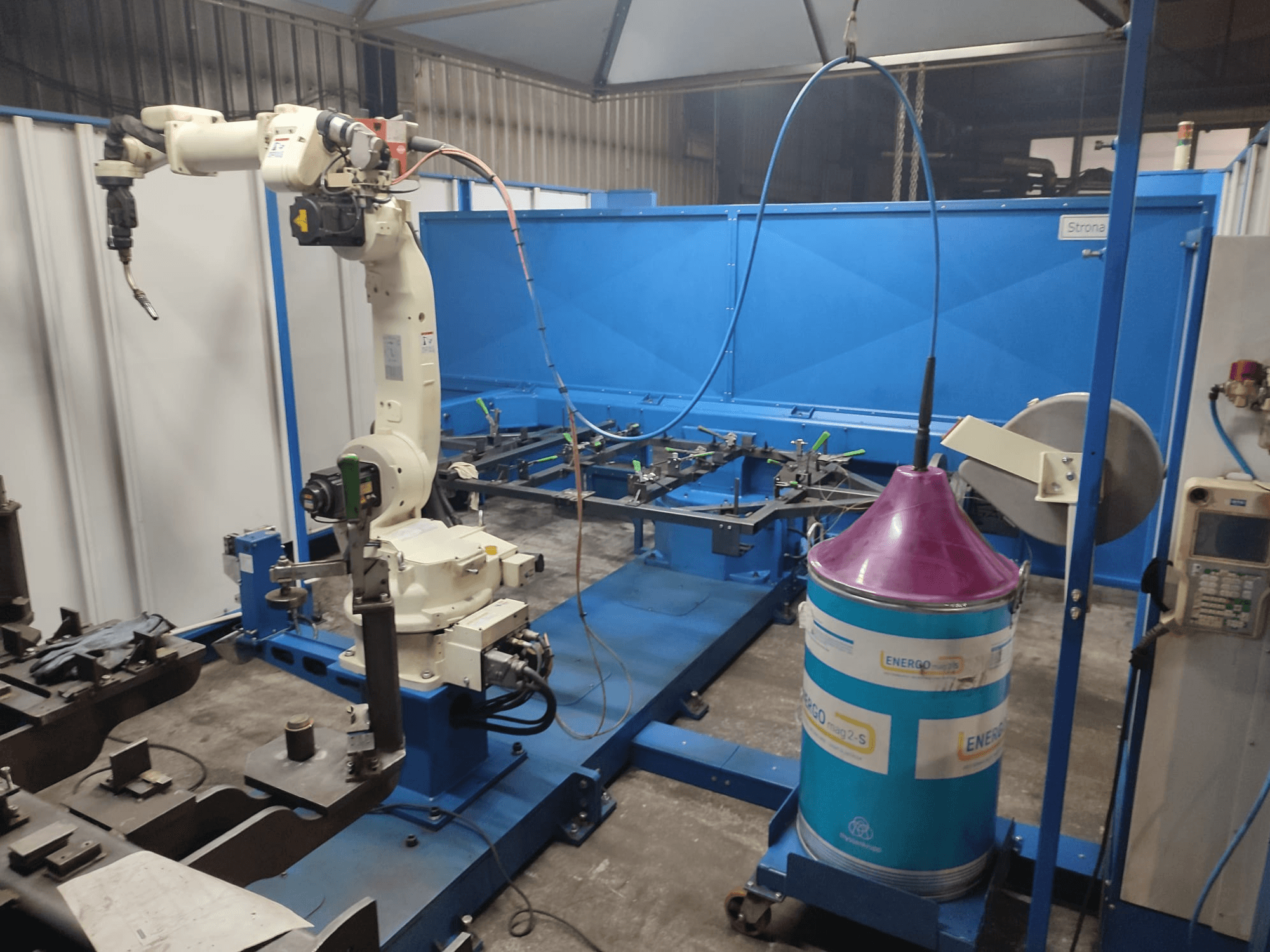 Pramoninė roboto ranka su baltos ir mėlynos spalvos pagrindu, įrengta dirbtuvėse su mėlynu darbo paviršiumi ir metaliniais komponentais.