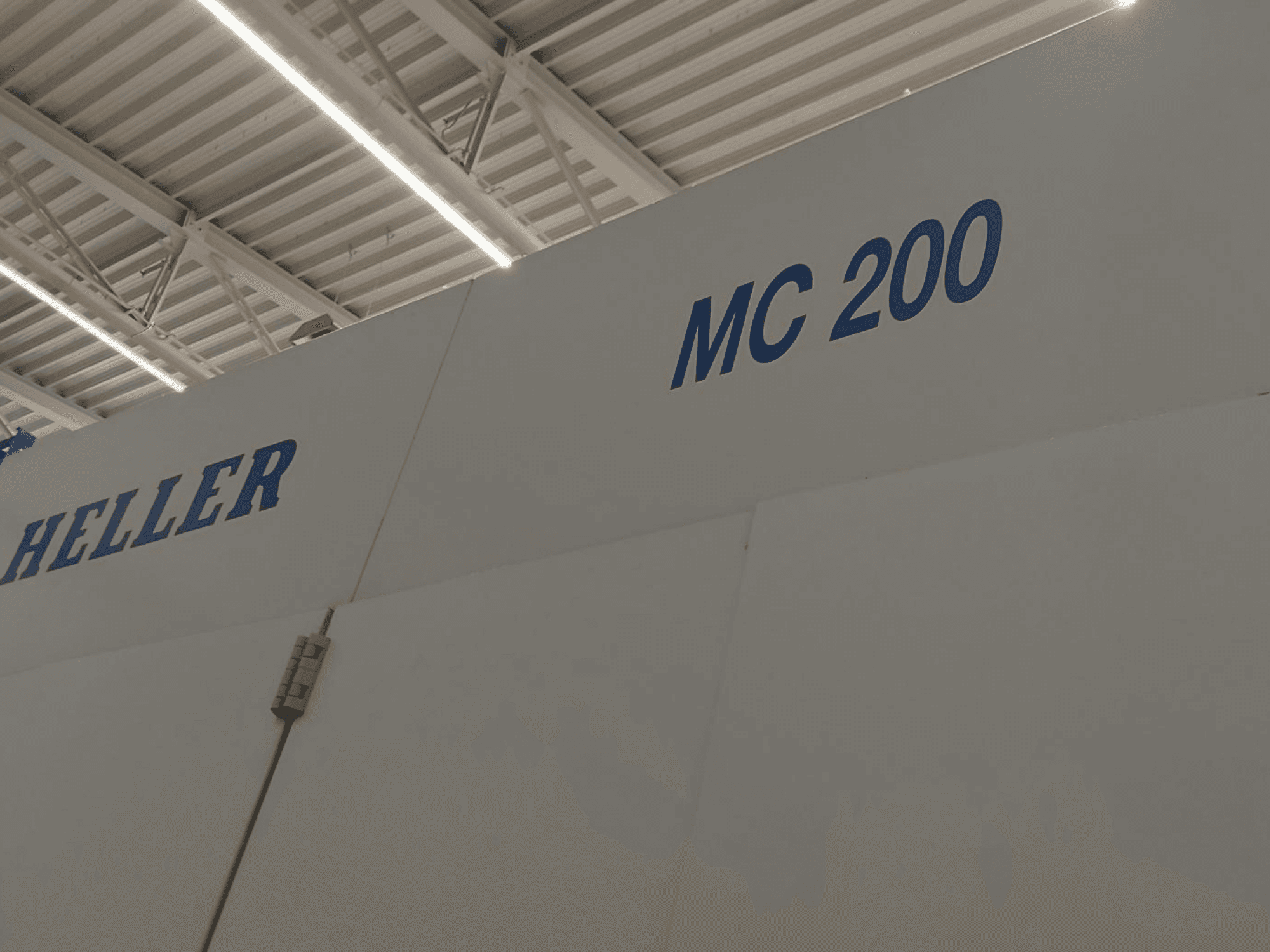 Horizontalusis apdirbimo centras "Heller MC 200", parodytas iš šono, ant balto paviršiaus mėlynais užrašais išryškintas prekės ženklas ir modelis.