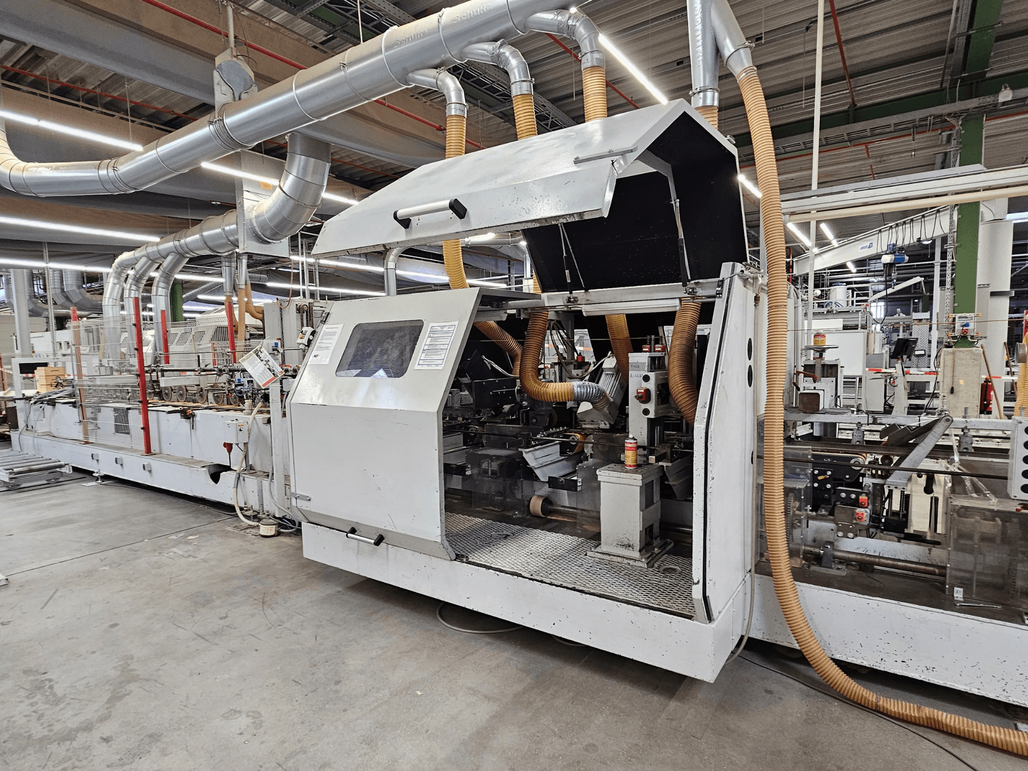 HOMAG FG63 briaunų šlifavimo staklės gamykloje, šoninis vaizdas, kuriame matyti darbiniai komponentai ir dulkių ištraukimo sistema.