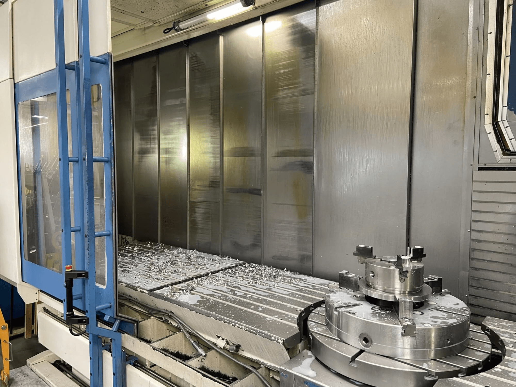 Vertikalus apdirbimo centras „AXA VHC-3-3000-M/E“, vaizdas iš priekio, kuriame matyti frezuotos metalo drožlės ant darbo stalo ir pjovimo įrankiai.