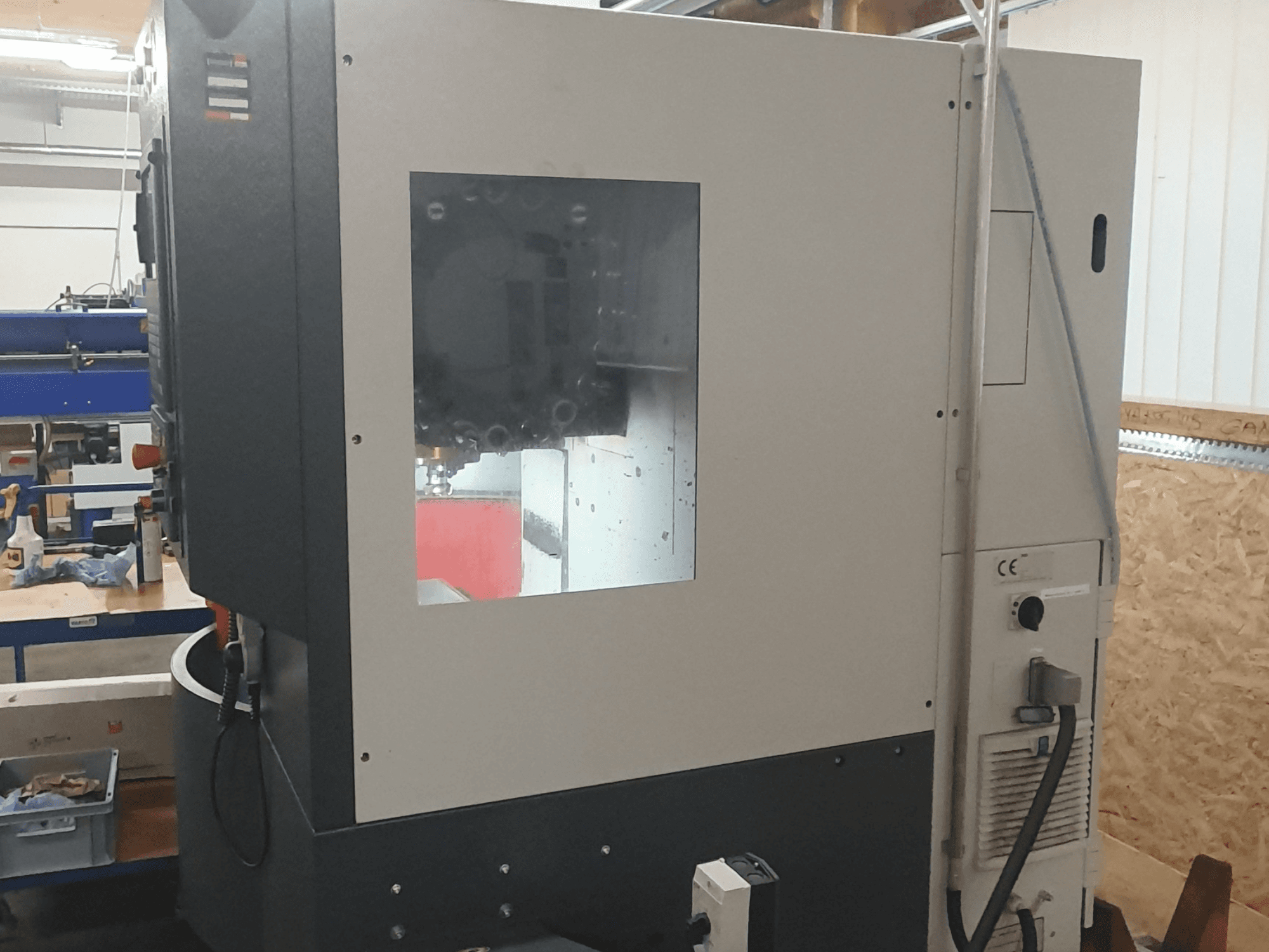 Metalo apdirbimo staklių vaizdas iš šono su matomu valdymo skydeliu, dideliu stebėjimo langu ir sudėtingais vidiniais komponentais.