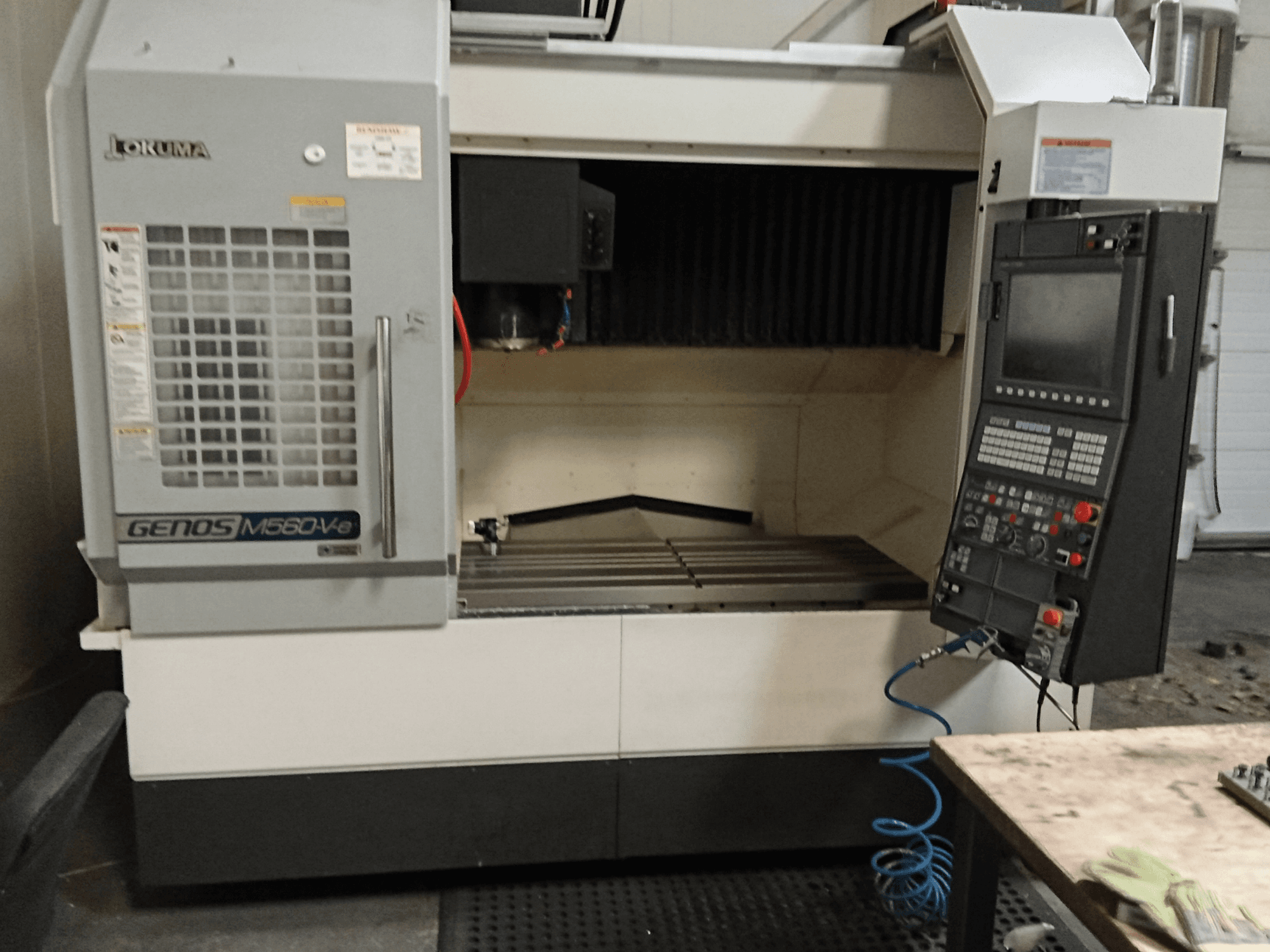 "Okuma GENOS M560-V", vertikalaus apdirbimo centro vaizdas iš priekio su valdymo pultu, CNC sąsaja ir matoma pjovimo zona.