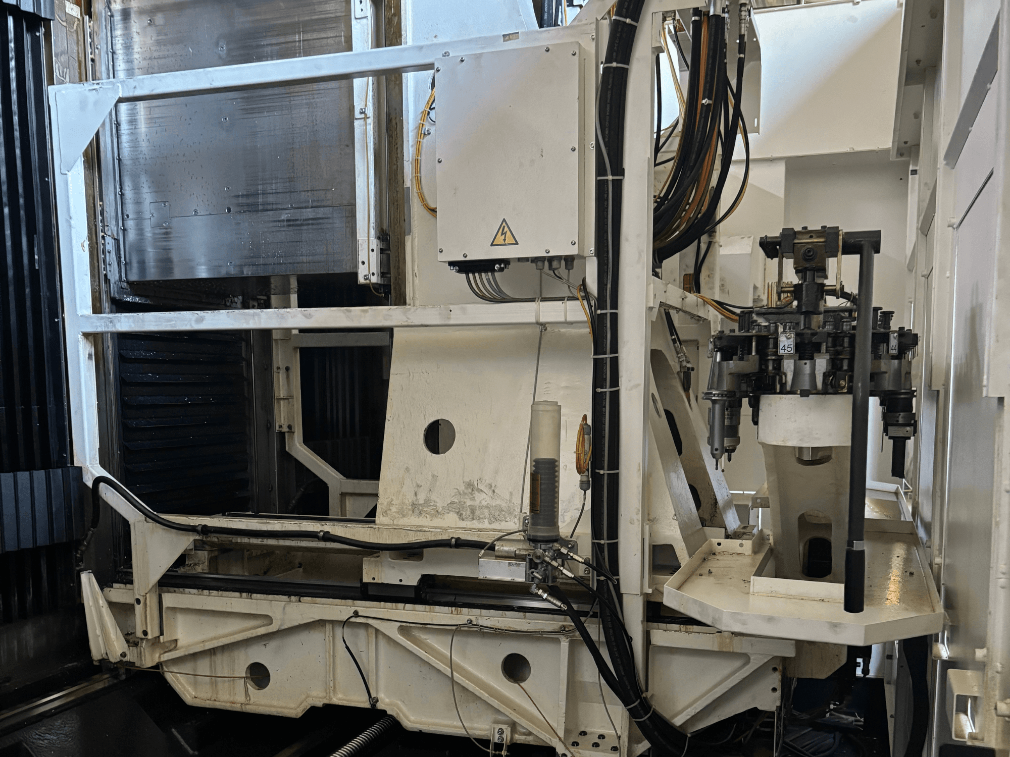 Pramoninių staklių šoninis vaizdas, kuriame matomos baltos CNC frezavimo staklės su matomais įrankiais, vamzdžiais ir valdymo skydeliu.