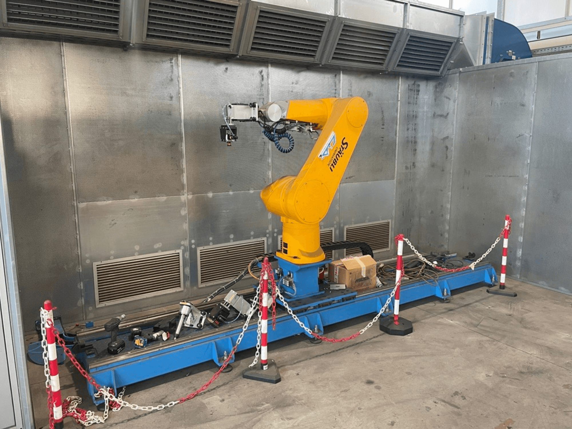 STAUBLI CS8C-RX160 roboto ranka dirbtuvėse, vaizdas iš šono, su kabeliais ir įvairiais įrankiais ant mėlyno pagrindo, apsupto apsauginiais barjerais.