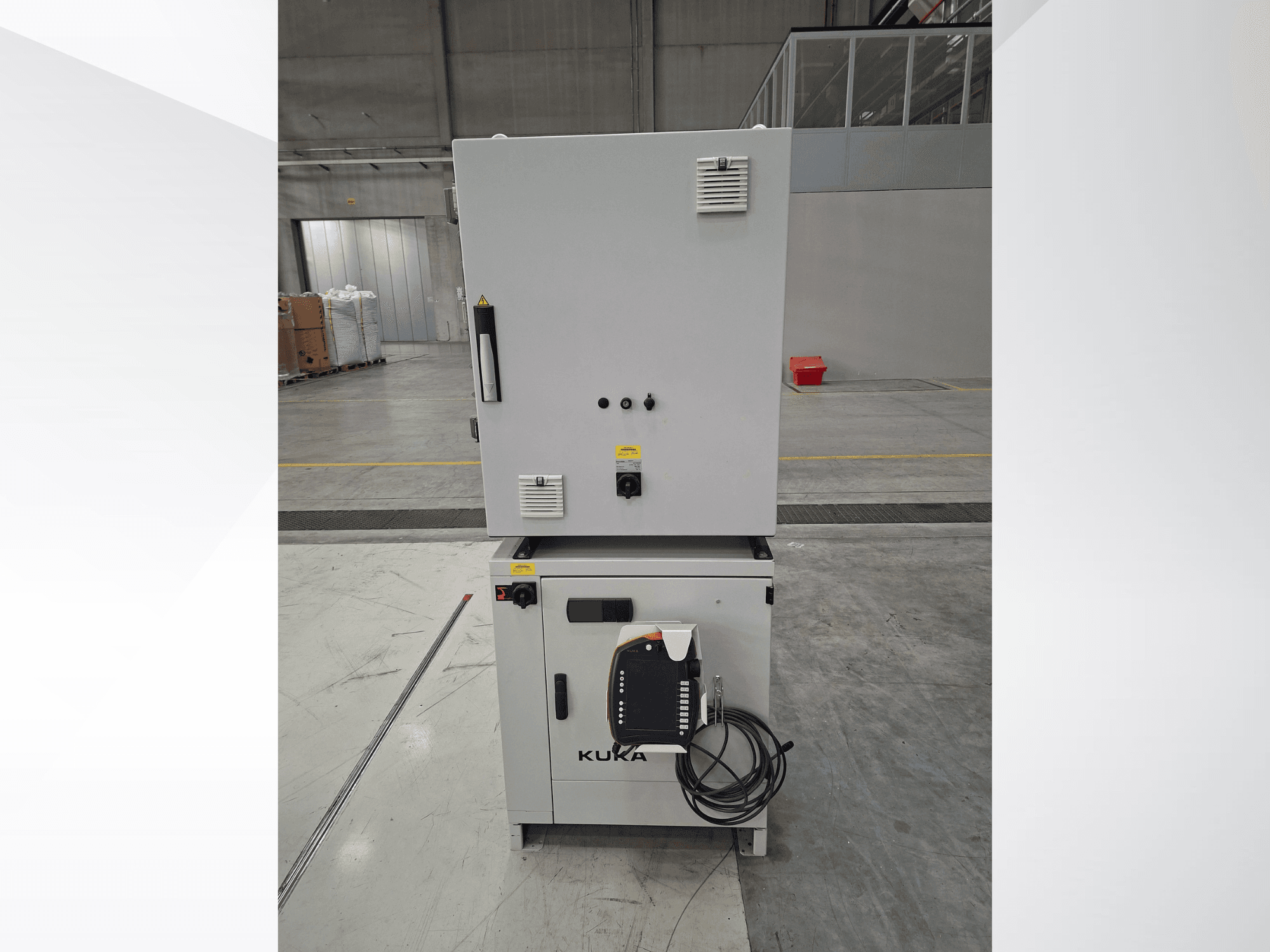 „KUKA KR 90 R2700 PRO C4 FLR“ robotinės rankos valdiklis, vaizdas iš priekio, kuriame matomas valdymo skydas ir saugos etiketės erdviame sandėlyje.