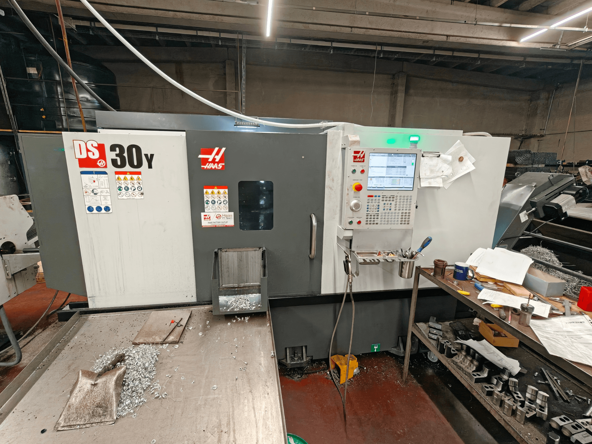 HAAS DS30Y horizontalus tekinimo staklės dirbtuvėse; vaizdas iš priekio, kuriame matomas valdymo pultas ir ant paviršiaus esančios metalo drožlės.