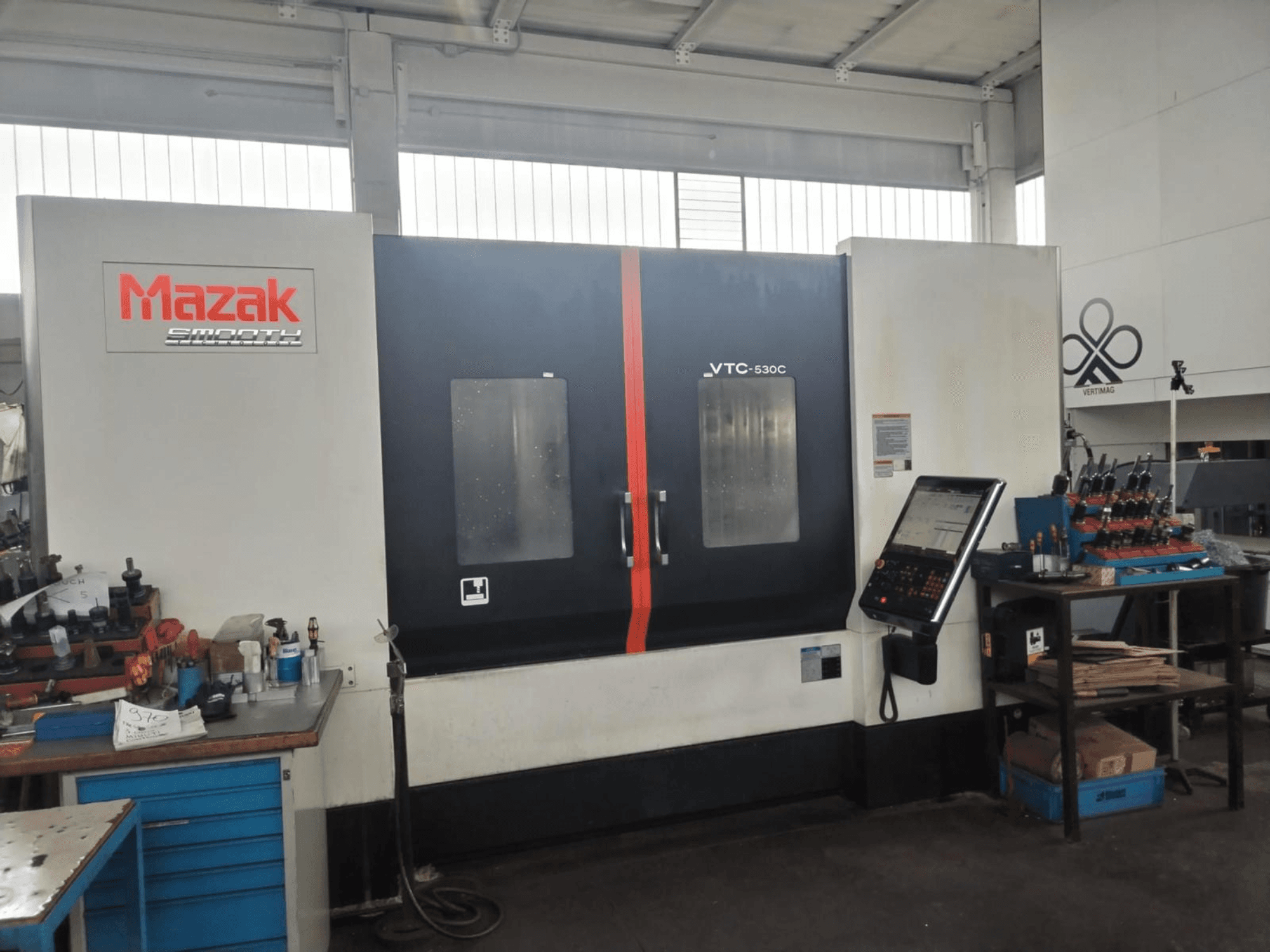 Dirbtuvėse esantis vertikalusis apdirbimo centras „Mazak VTC-530C“ su valdymo pultu ir įrankiais, išdėstytais ant šoninio stalo.