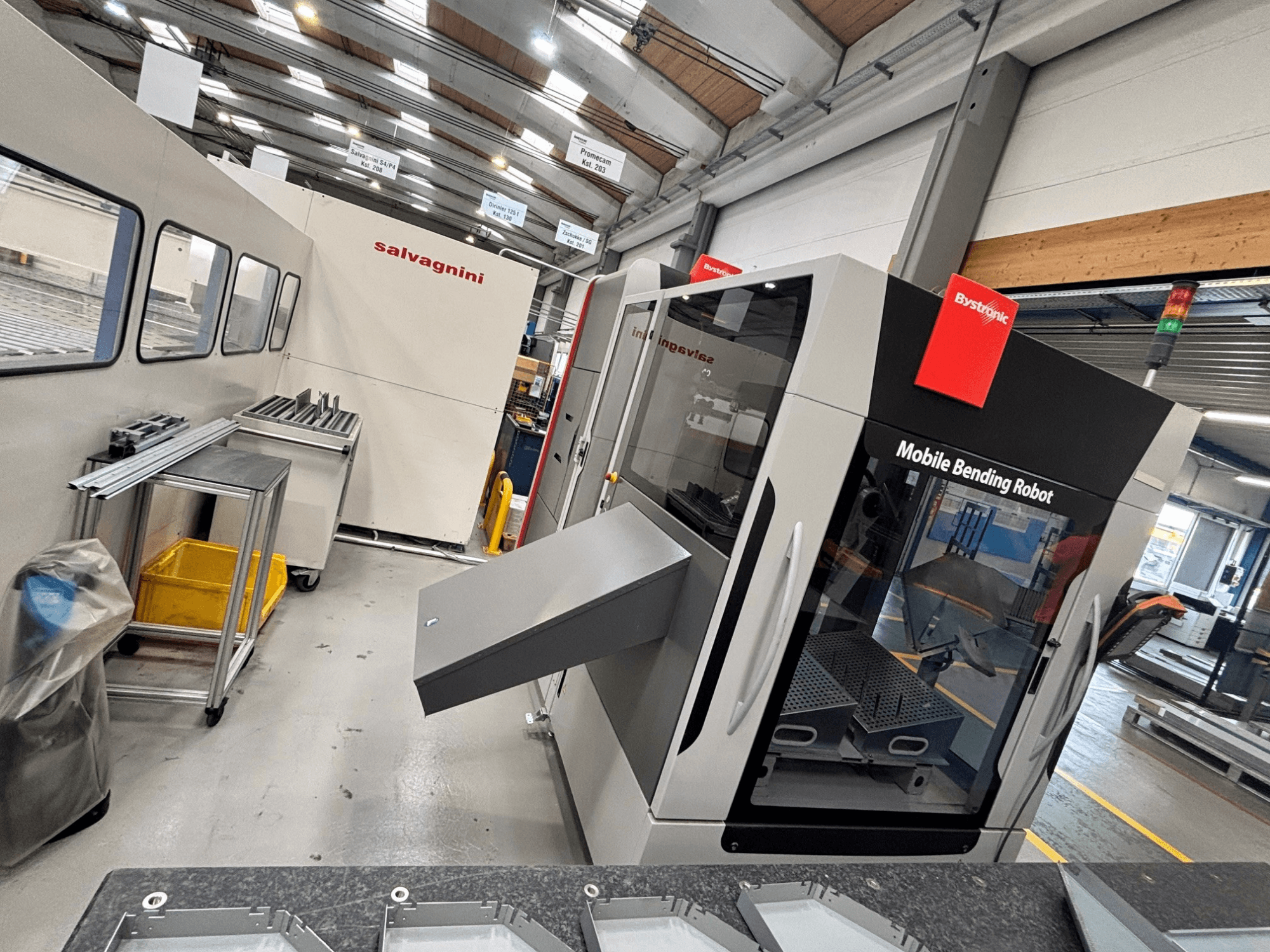 „Bystronic Xpert 40/1030“ presinis lenkimo staklės, vaizdas iš priekio, kuriame matomas judamasis lenkimo robotas ir darbo zona su metalinėmis detalėmis ant stalo.