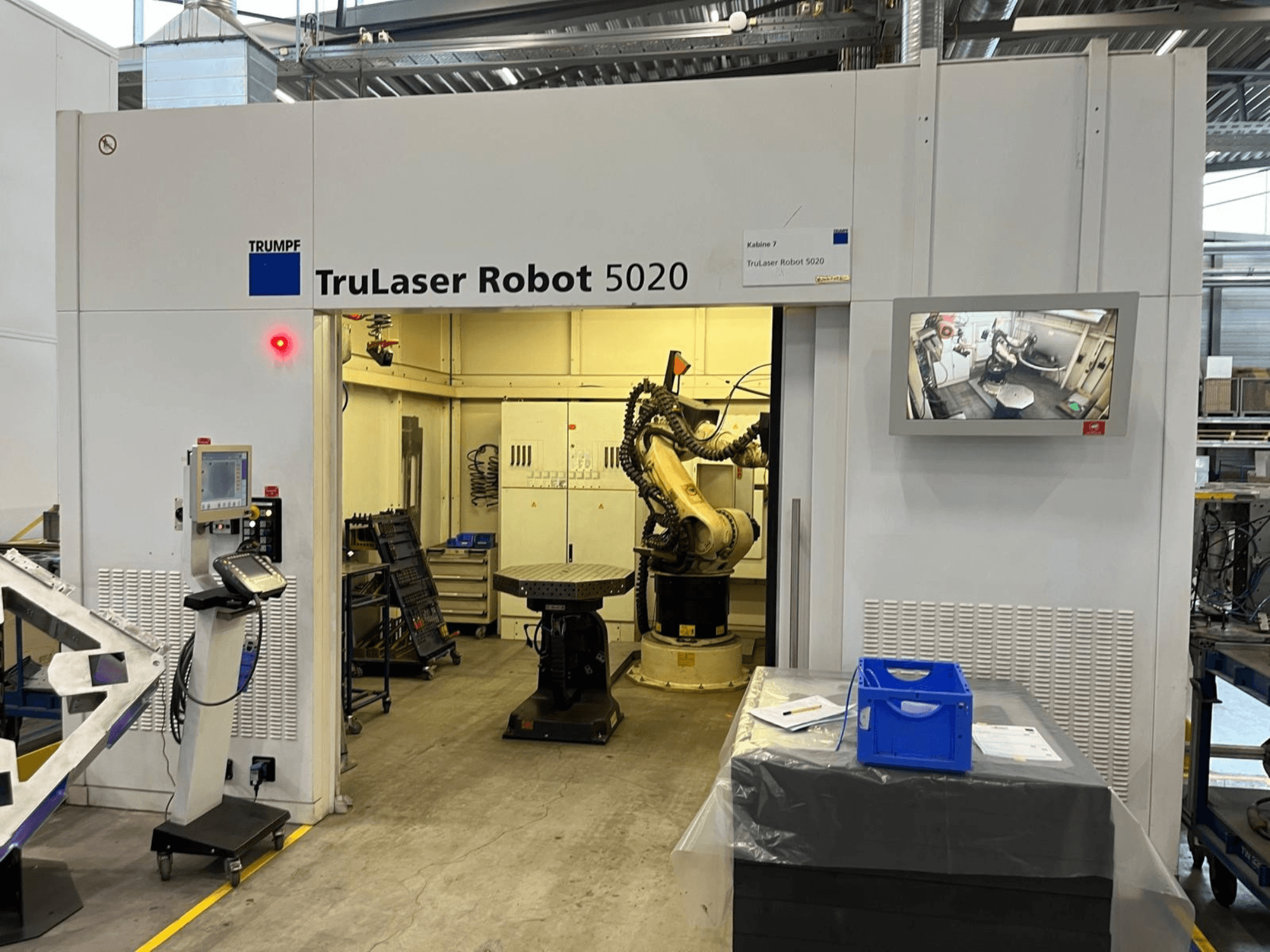 „TRUMPF TruLaser Robot 5020“ pramoninėje aplinkoje: matomi valdymo elementai, robotinė ranka ir operacijas rodantis monitorius.