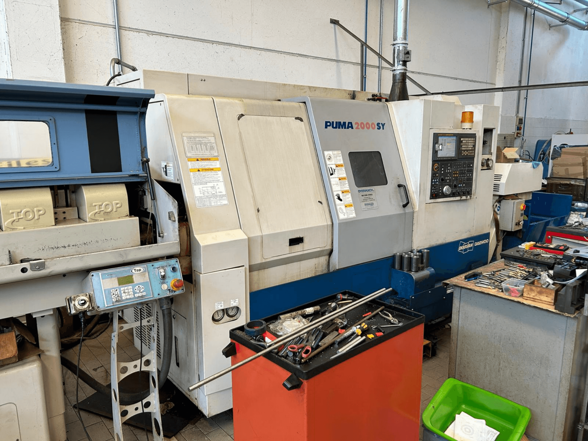 „DOOSAN PUMA 2000SY“ horizontalioji tekinimo staklė dirbtuvėse, vaizdas iš šono, kuriame matomas valdymo pultas ir šalia esantys įrankiai.