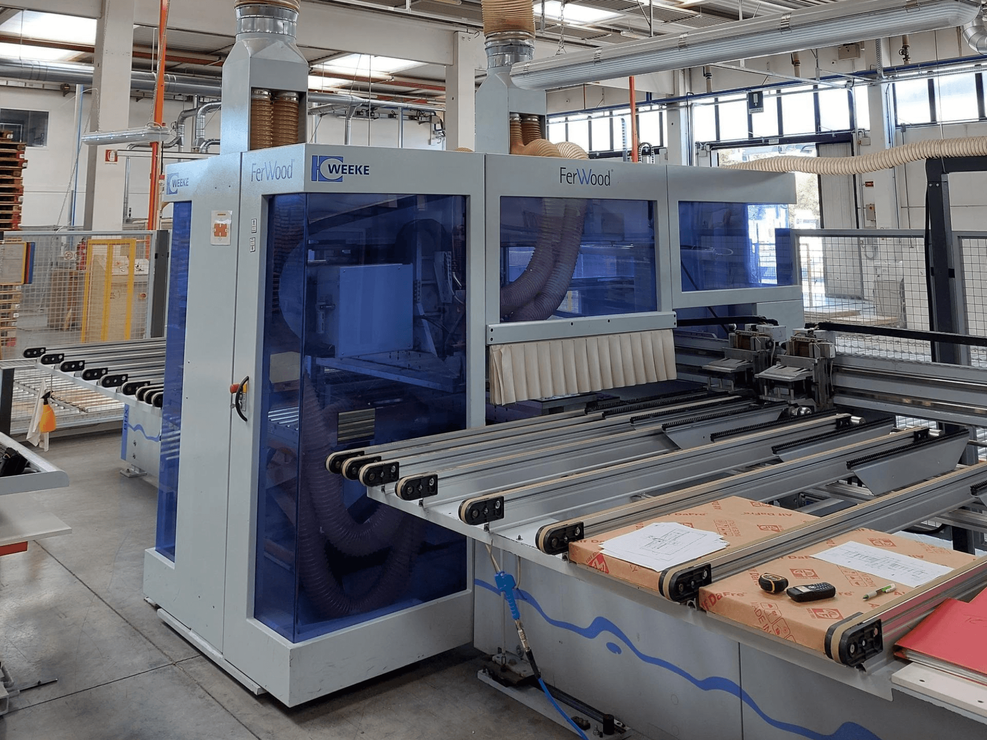 Weeke FerWood CNC staklės dirbtuvėse, vaizdas iš priekio, kuriame matyti mėlynai nuspalvinti skydeliai, valdymo mygtukai ir darbo stalas su medžiagomis.