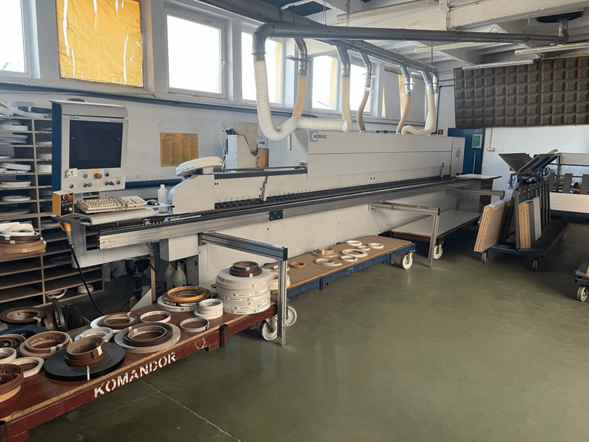 "HOMAG" kraštų apklijavimo mašina dirbtuvėse, su valdikliais ir įvairiomis kraštų apklijavimo medžiagomis ant mėlyno vežimėlio. Vaizdas iš šono.