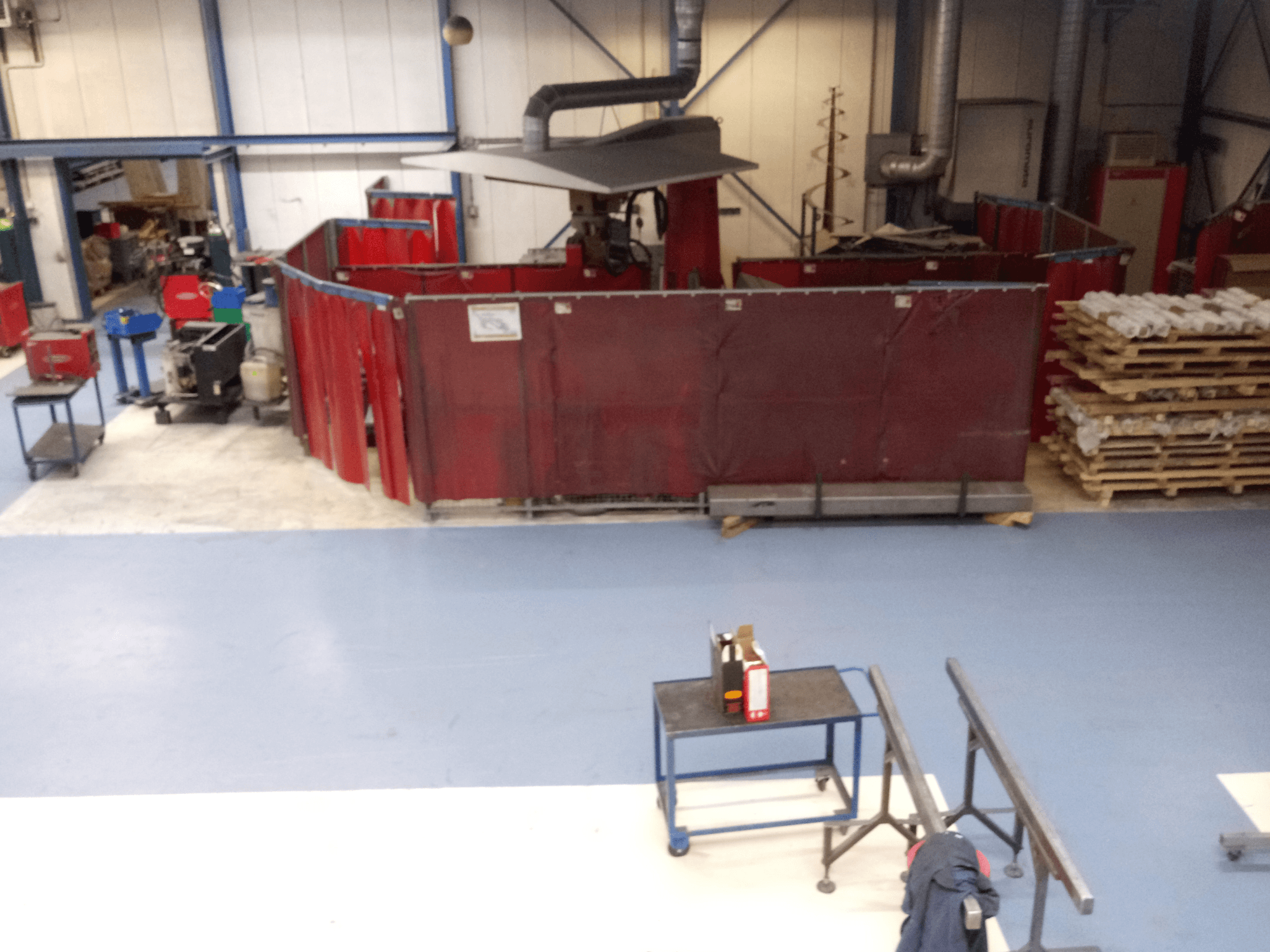 Pramoninė darbo vieta su raudonomis suvirinimo užuolaidomis, mašinomis ir padėklais; vaizdas iš viršaus, rodantis išdėstymą ir įrangą.