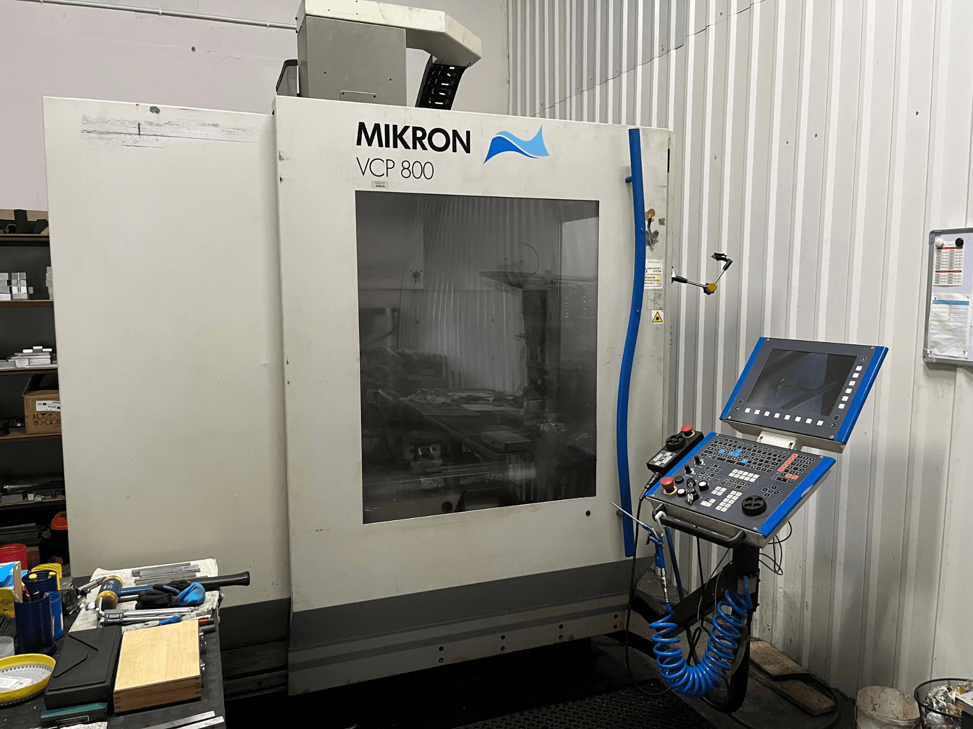 Vertikalusis apdirbimo centras „MIKRON VCP 800“, vaizdas iš priekio, su valdymo pultu ir įrankiais ant darbastalio pirmame plane.