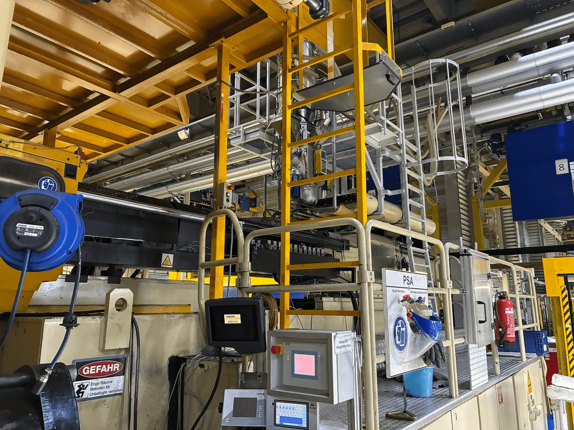 Pramoninių mašinų valdymo skydas su mėlyna saugos įranga ir geltonais pastoliais, vaizdas iš šono, kuriame matyti vamzdžiai ir įspėjamieji ženklai.