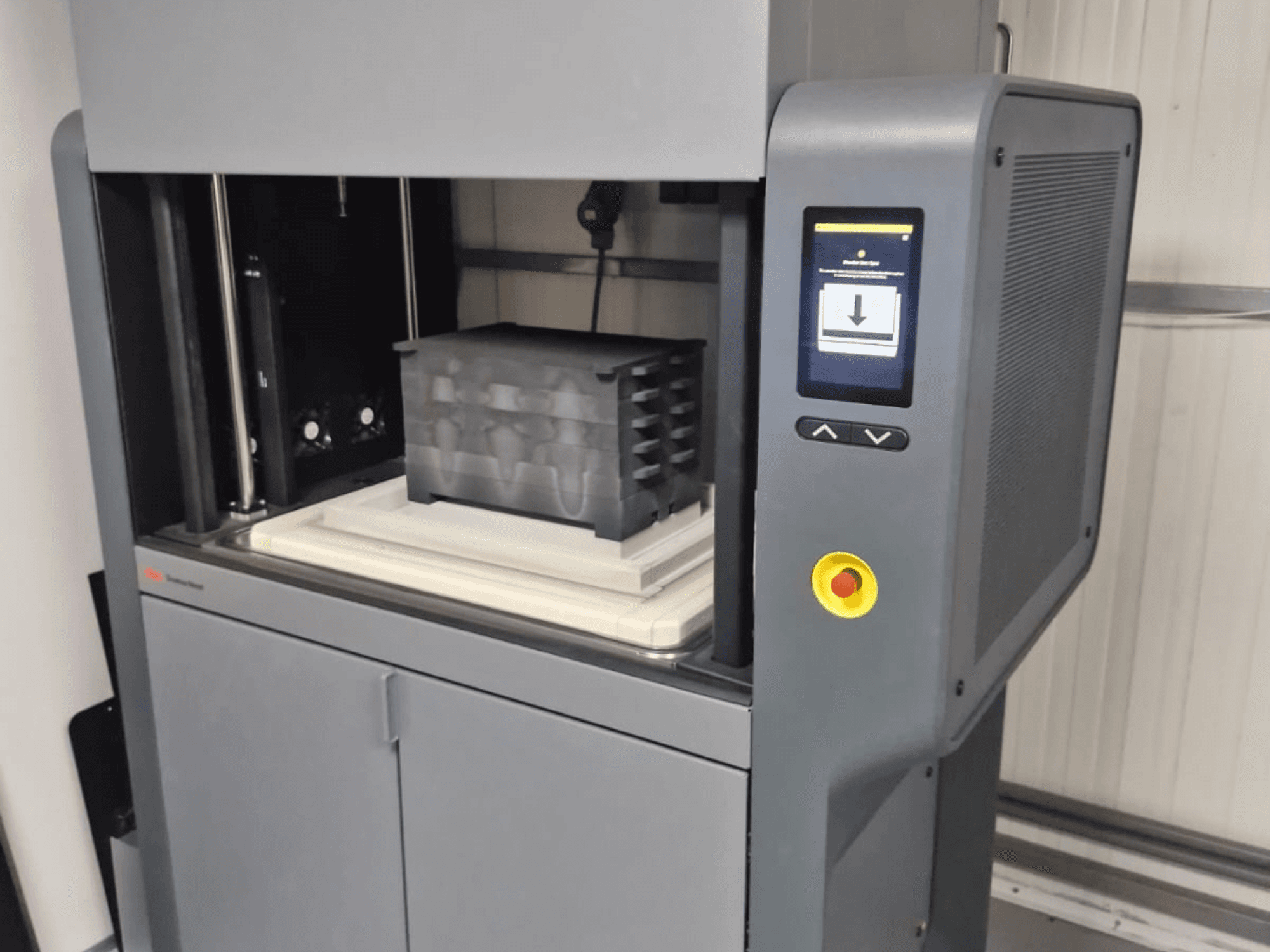 "Desktop Metal" 3D spausdintuvas "Metal Studio 2", priekinis vaizdas, kuriame matyti valdymo skydelis ir spausdinimo lova su juodu konteineriu.