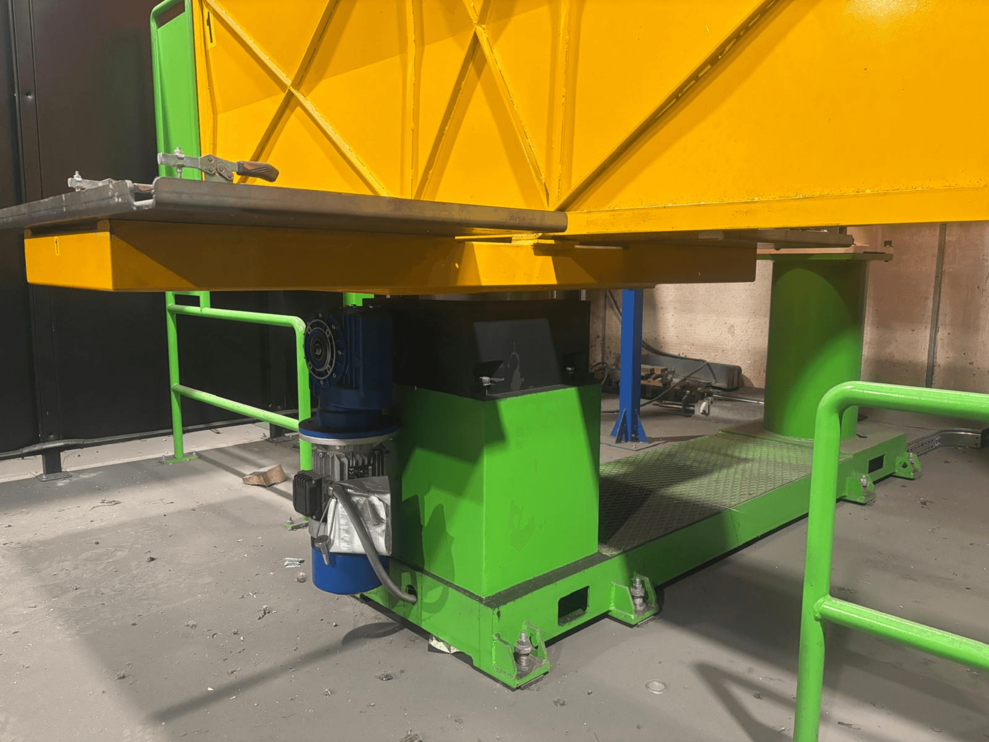 Žalios ir geltonos spalvos pramoninės mašinos iš žemo priekinio žvilgsnio su varikliu ir platforma su tekstūruotu paviršiumi.