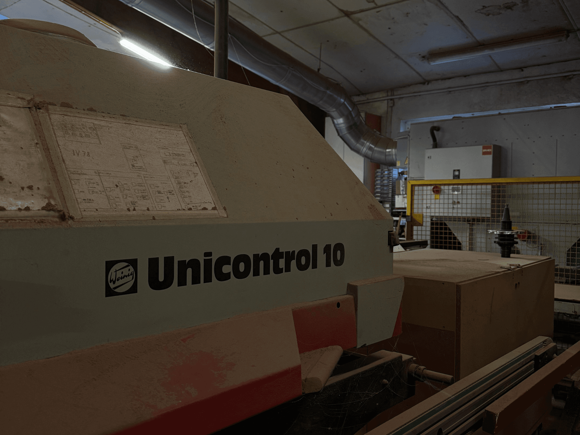 Mašina "Weinig Unicontrol 10", žiūrint iš šono, dulkėtas paviršius, ant priekinio skydelio matomos techninės schemos.