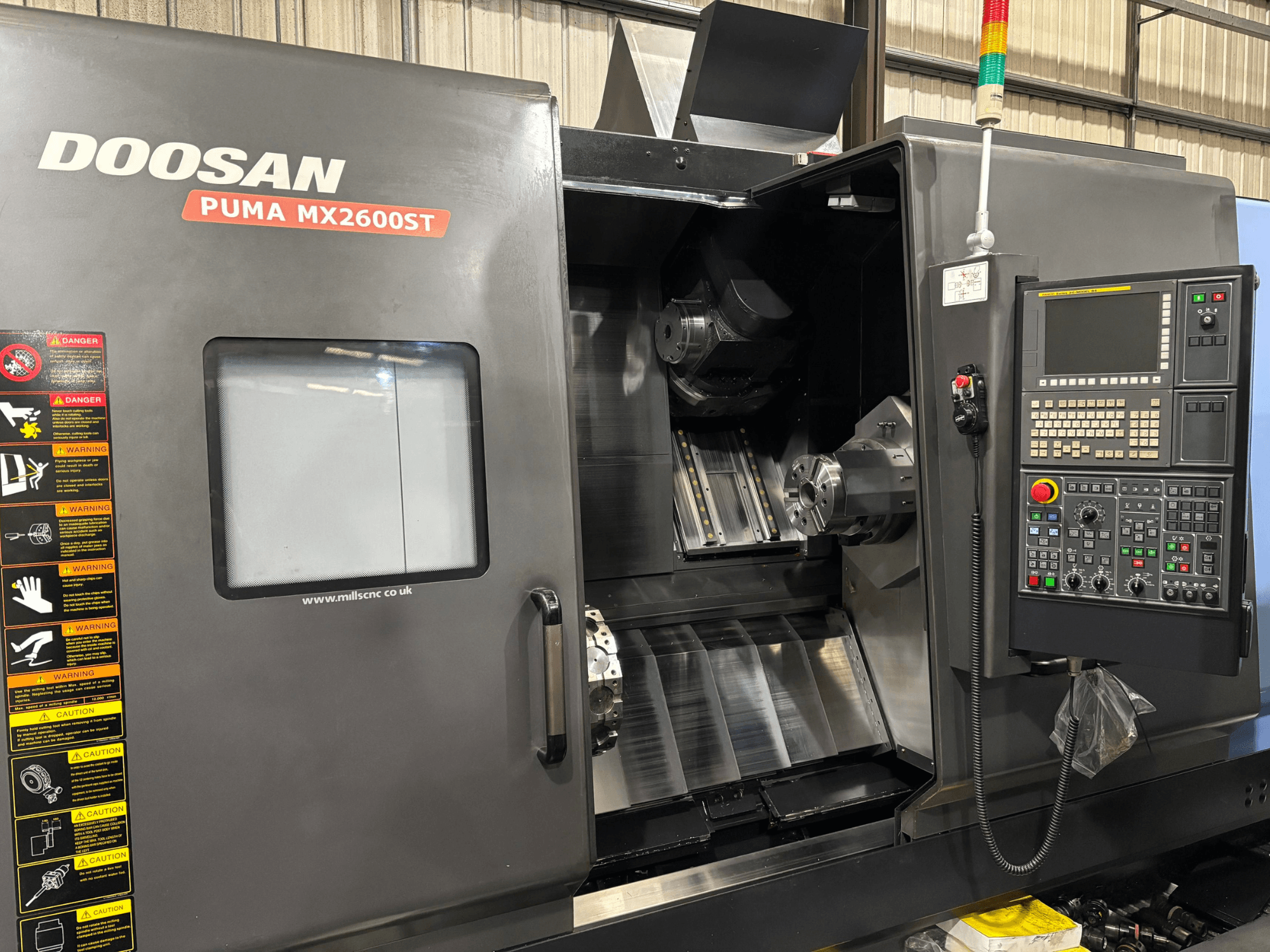 CNC staklės Doosan PUMA MX2600ST, priekinis vaizdas, kuriame matomas valdymo skydelis, suklys ir apdirbimo zona su matomais saugos įspėjimais.
