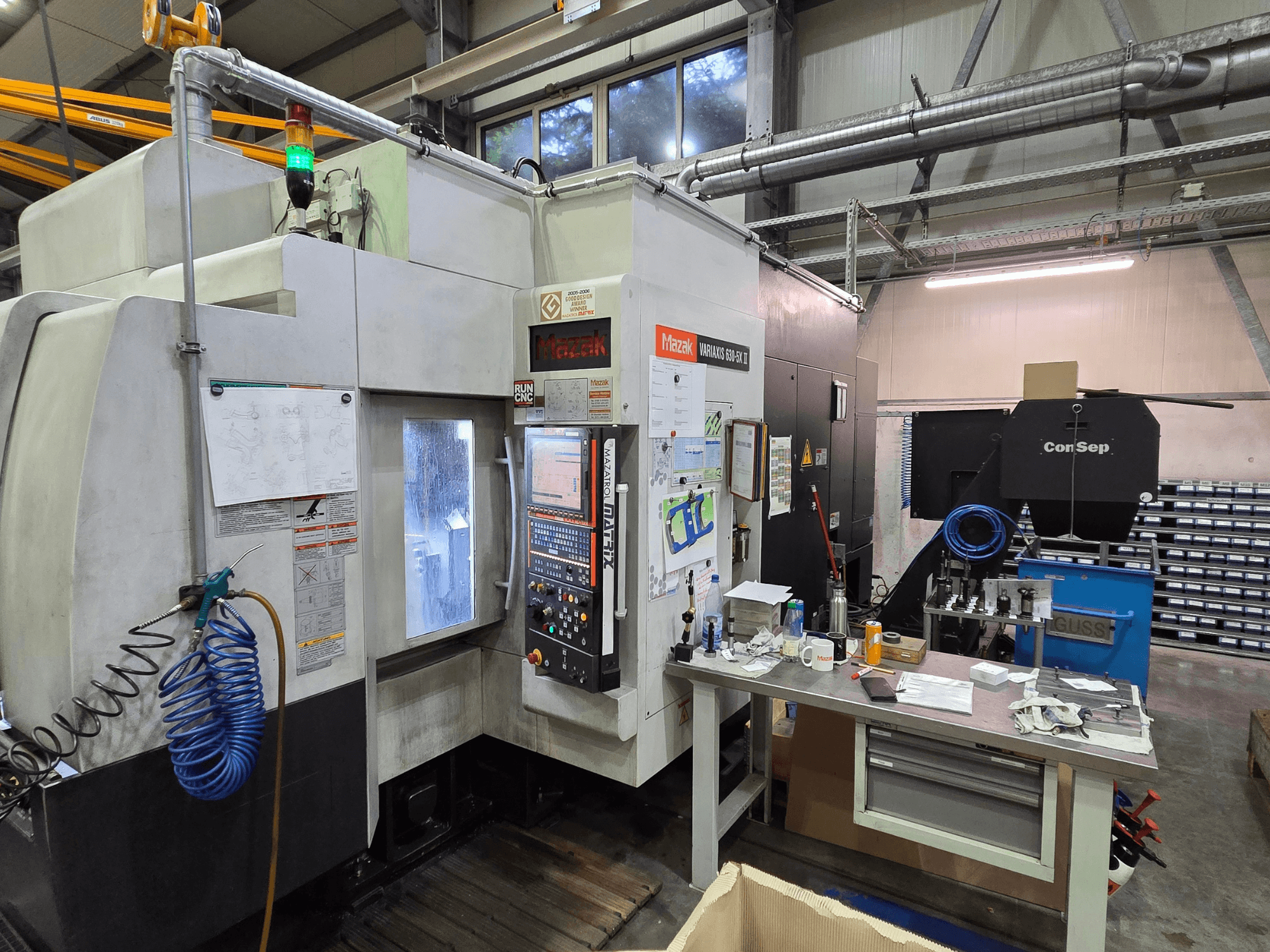 Mazak Variaxis 630-5X CNC staklės dirbtuvėse, vaizdas iš priekio su valdymo skydeliu, žarna ir priedais ant šoninio stalo.