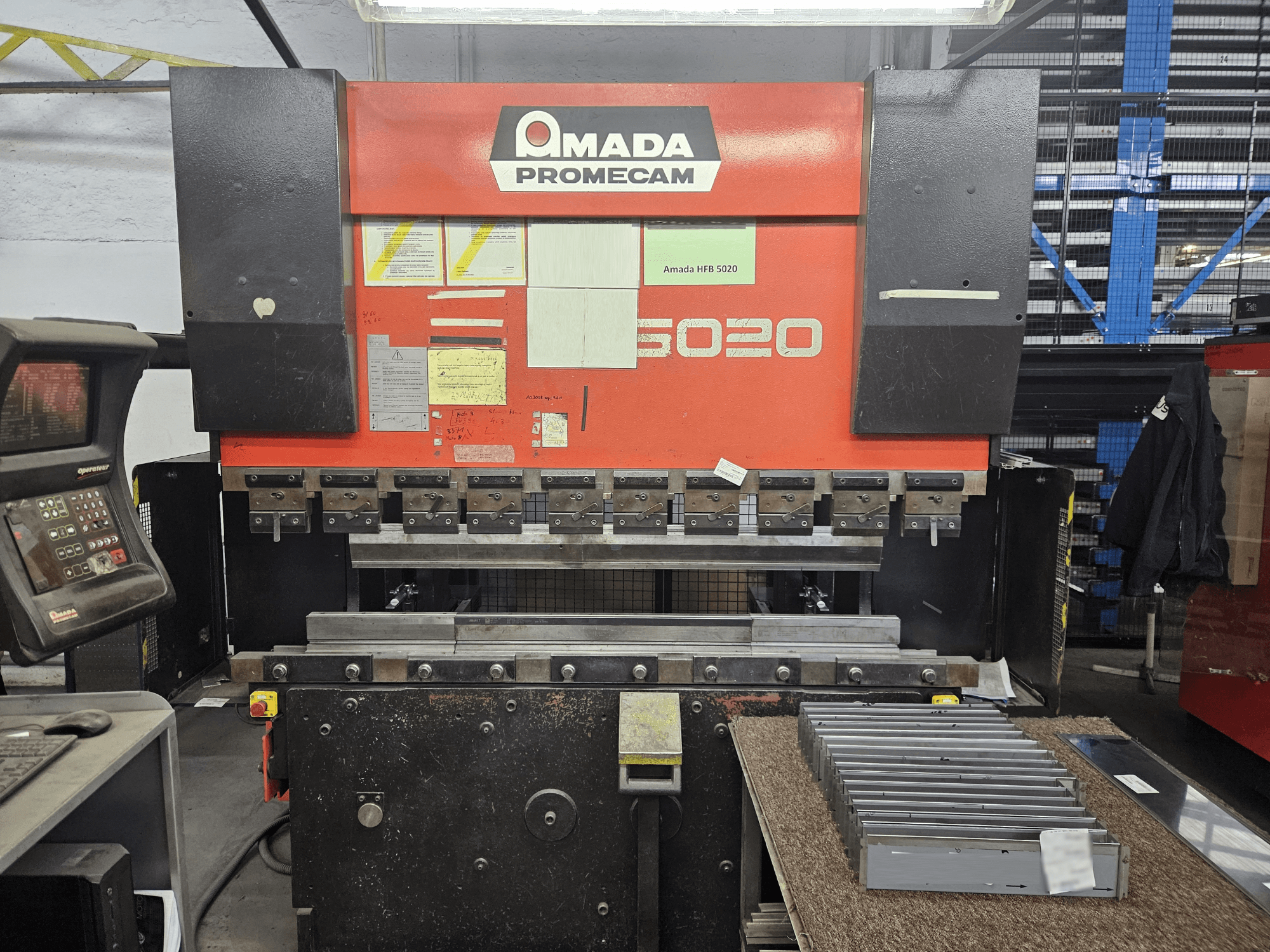 Amada HFBO 5020 preso stabdys iš priekio, su valdymo skydeliu ir ant darbinio paviršiaus sudėliotomis metalinėmis detalėmis.