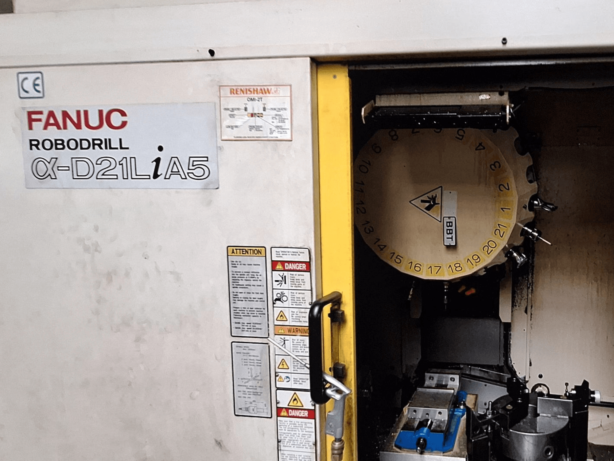 FANUC Robodrill α-D21LiA5, vaizdas iš priekio. Rodoma įrankių karuselė ir apdirbimo zona su spaustuvais ir įspėjimais ant durelių.