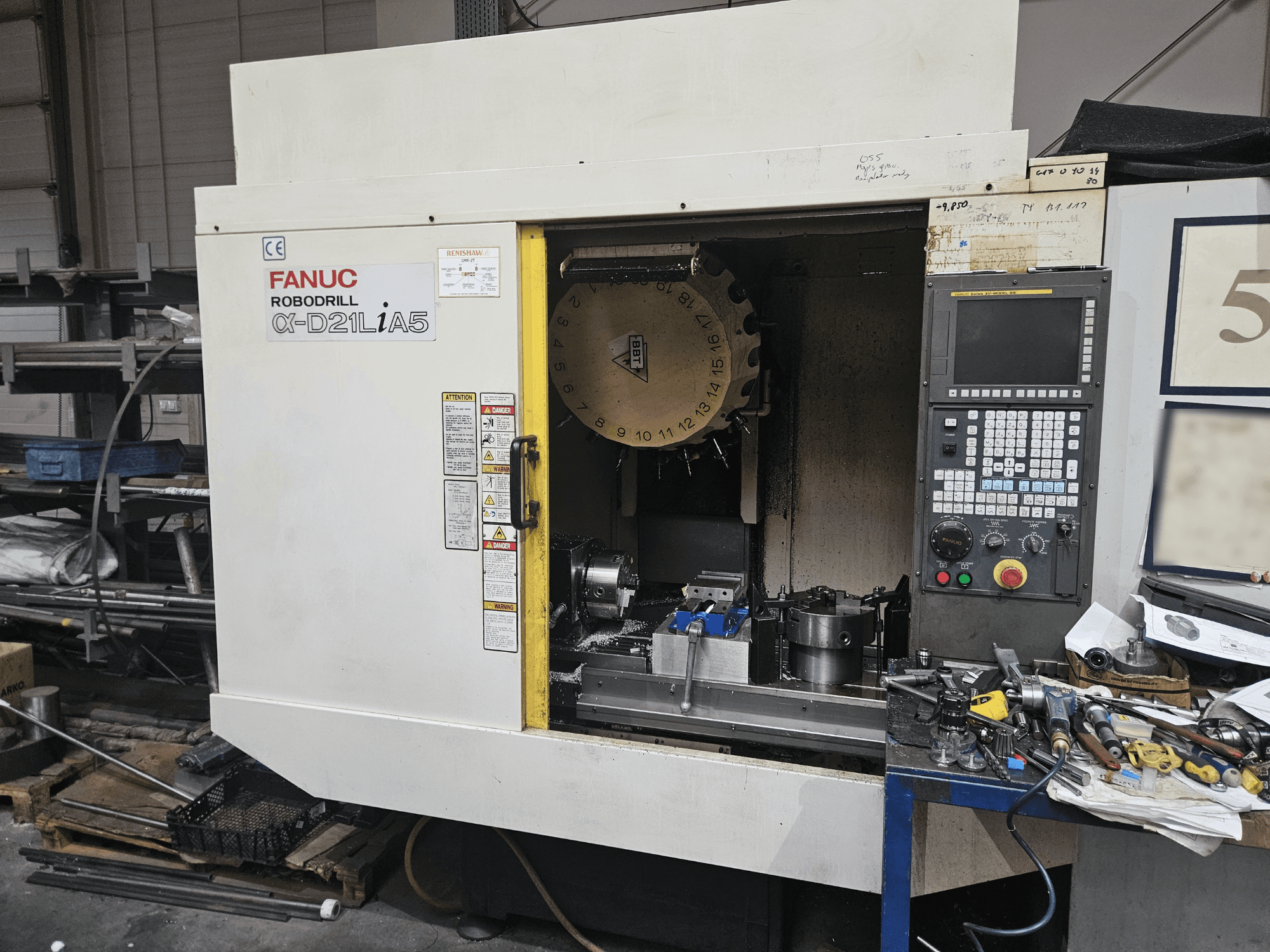 FANUC Robodrill α-D21LiA5, priekinis vaizdas, kuriame matomas įrankių laikiklis ir valdymo skydelis, apsuptas įrankių ir įrangos dirbtuvėse.