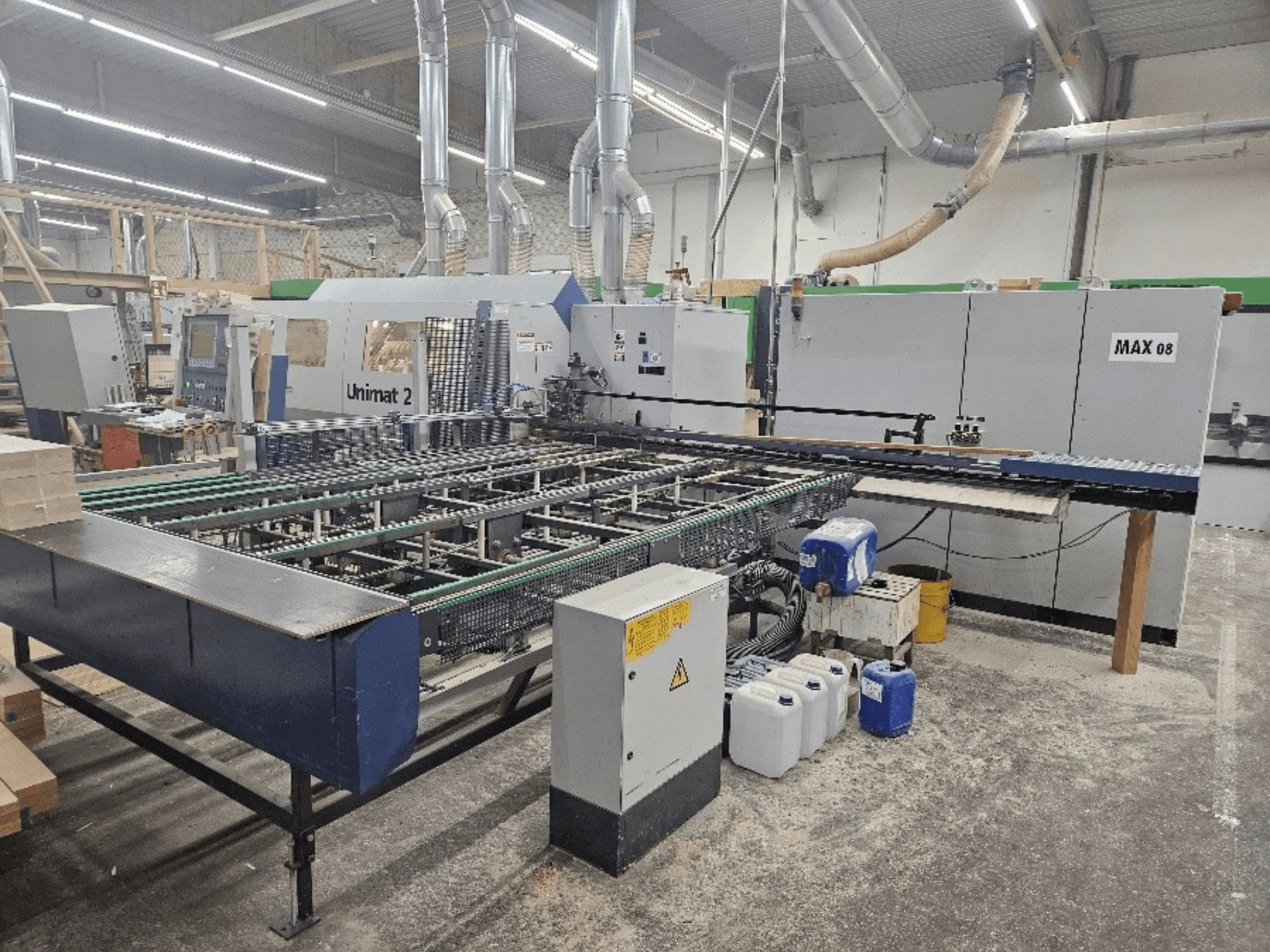 "Unimat 2 CNC" staklių vaizdas iš šono dirbtuvėse su konvejerių juostomis ir techniniais komponentais, matomas "Max 08" įrenginys.
