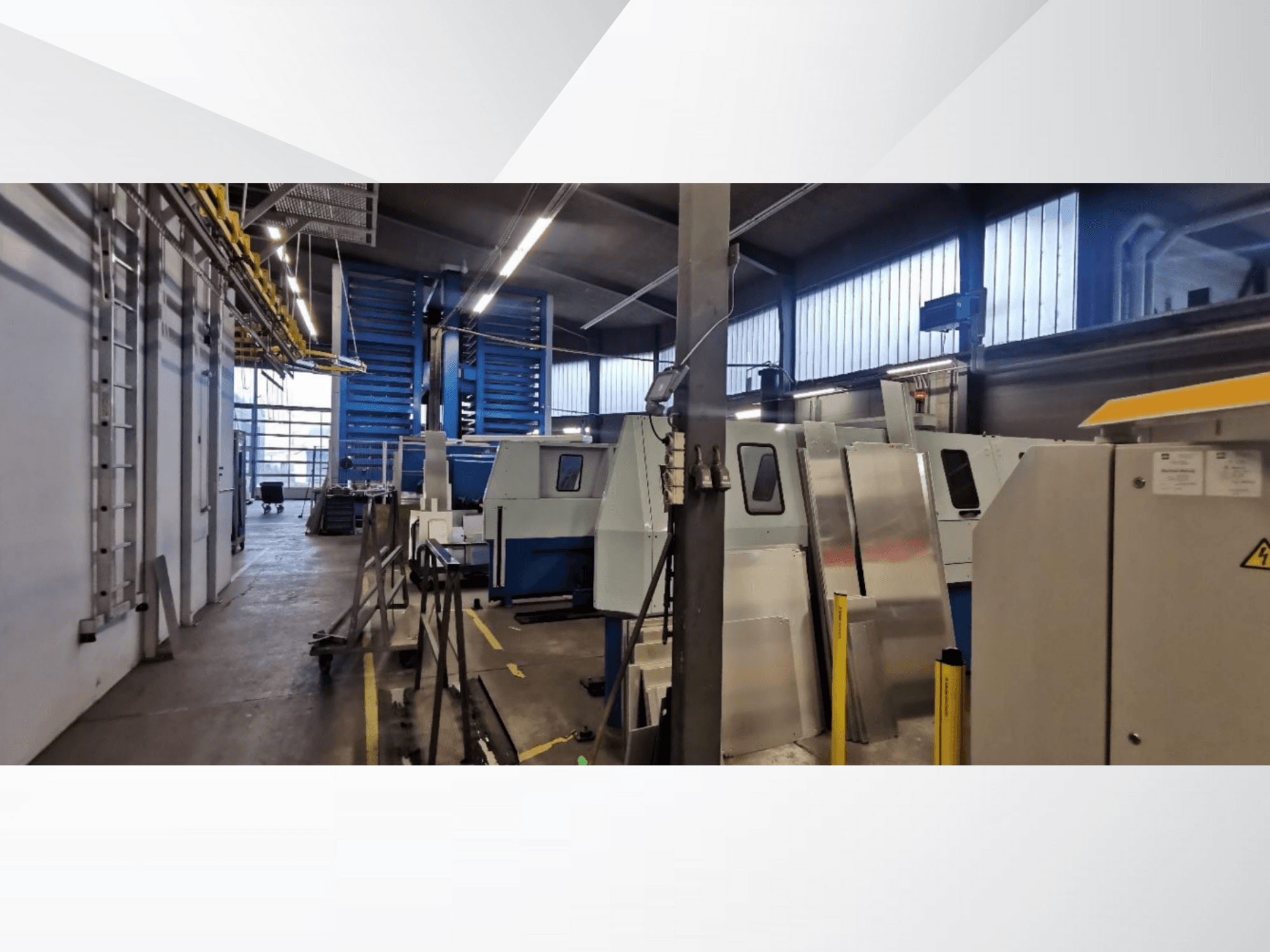 Pramoninių dirbtuvių scena su keliomis mašinomis ir įranga iš šono, su metaliniais paviršiais ir matomais saugos įtaisais.
