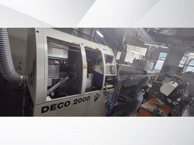 DECO 2000 CNC staklės dirbtuvėse, vaizdas iš priekio, matomi valdikliai ir ilgo ruošinio sąranka dešinėje.