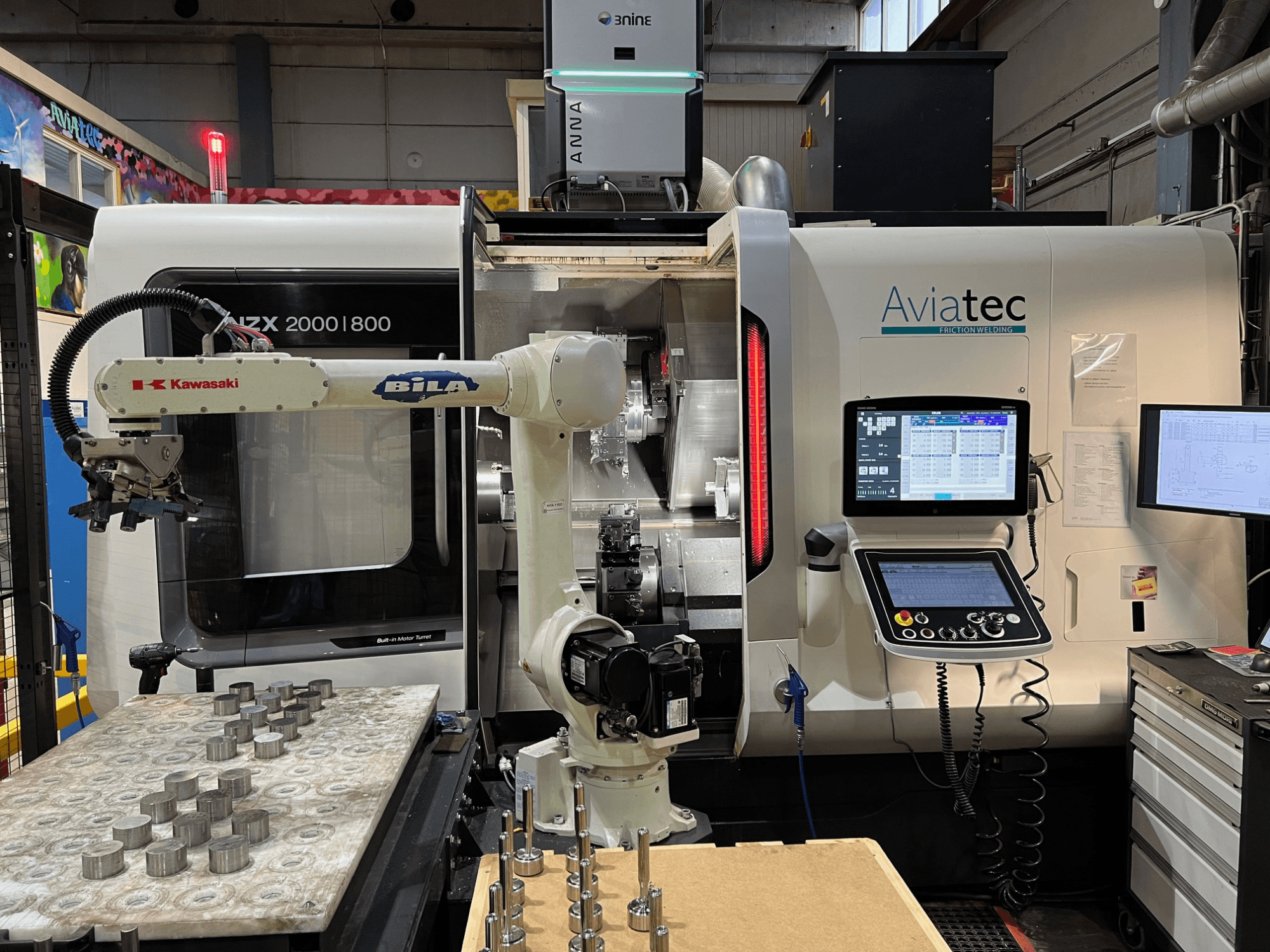 Horizontali tekinimo staklė „DMG MORI NZX 2000/800SY“ su „Kawasaki“ robotine ranka, valdymo pultu ir metalinėmis detalėmis ant darbo stalo.