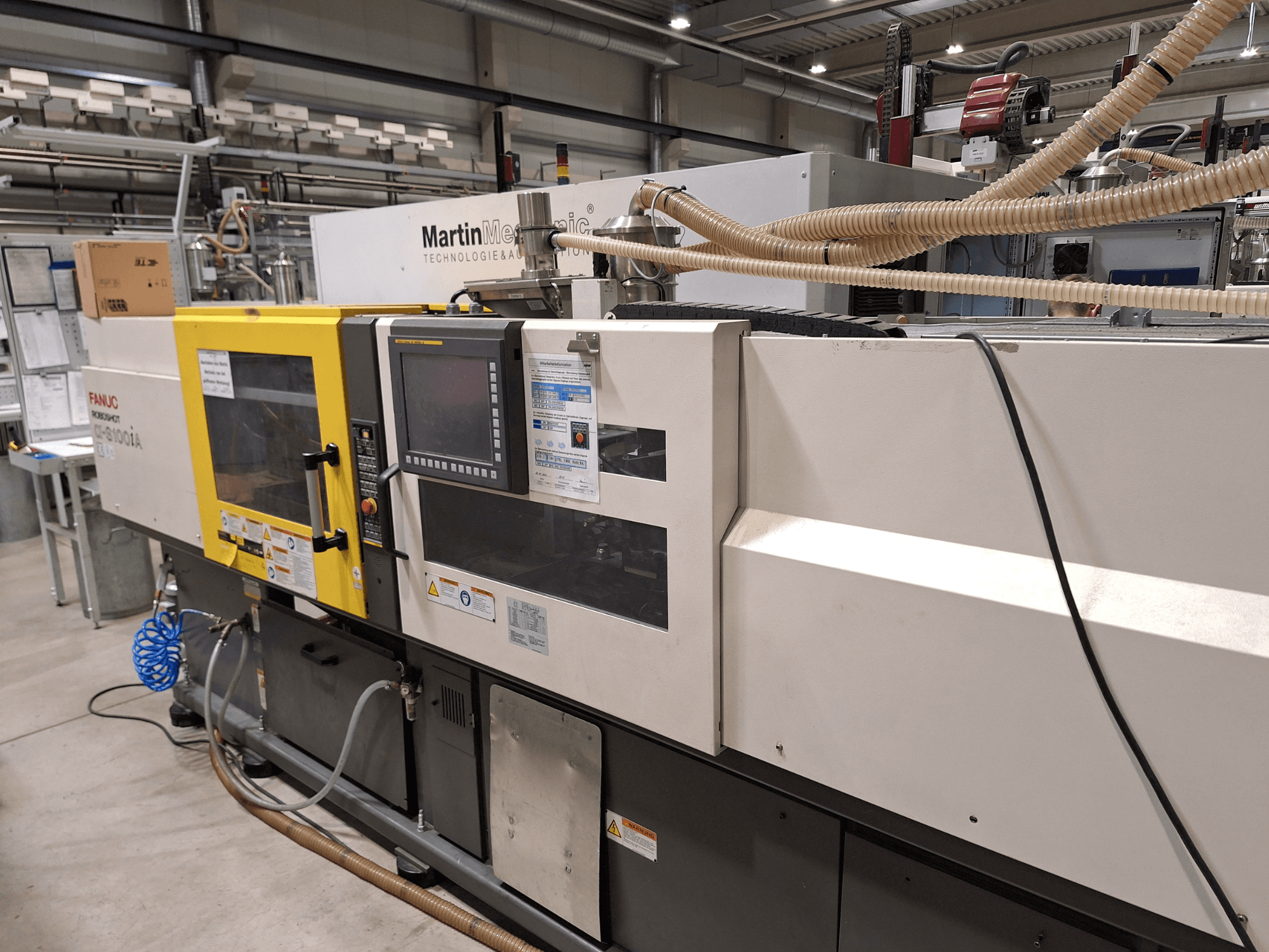 FANUC Roboshot S100iA elektrinė įpurškimo mašina, vaizdas iš šono, su jutikliniu ekranu ir saugos etiketėmis.