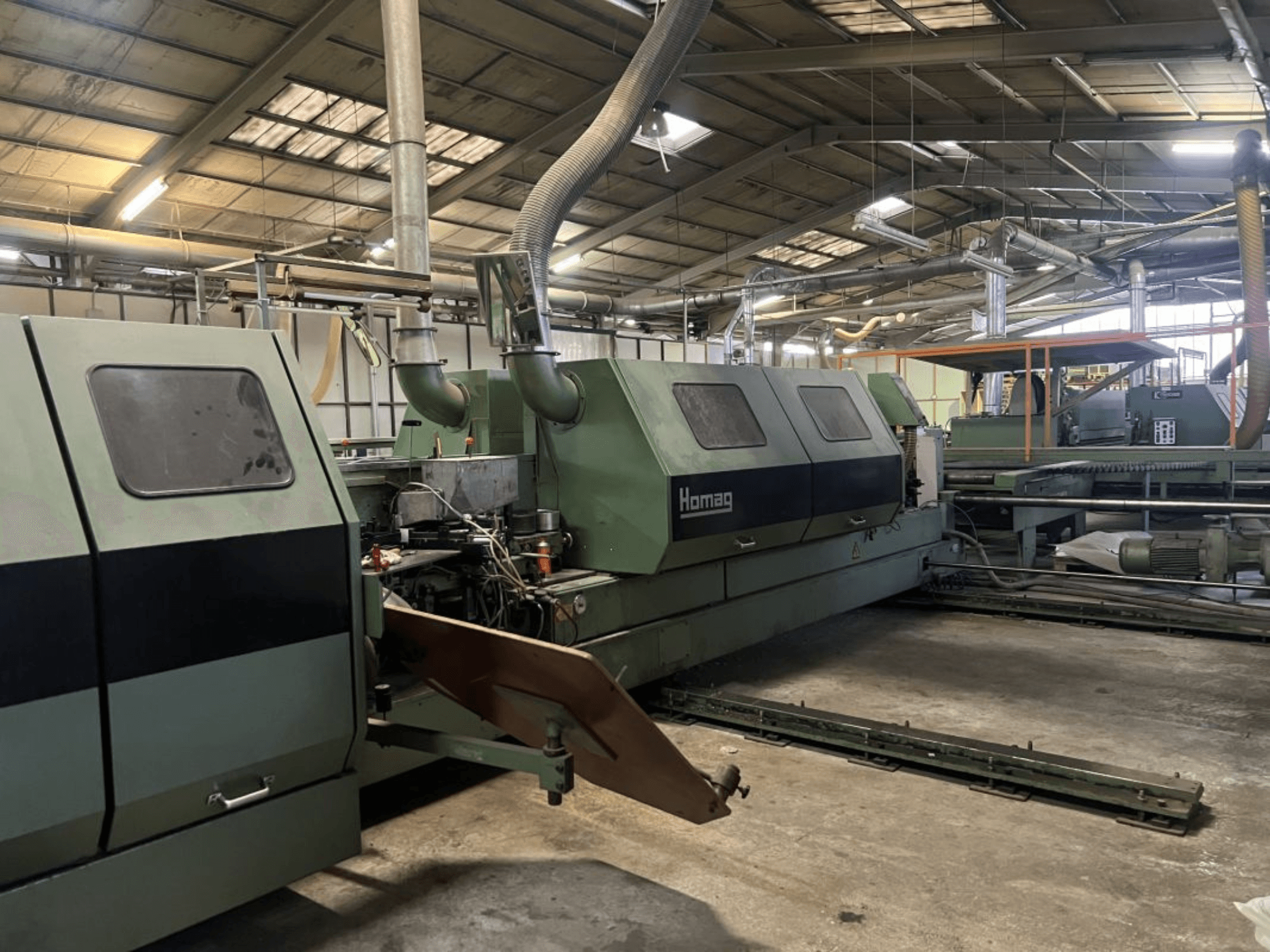 Žalios spalvos "Homag" pramoninė mašina, vaizdas iš šono, matomi veikiantys komponentai ir valdymo skydelis sandėlyje.