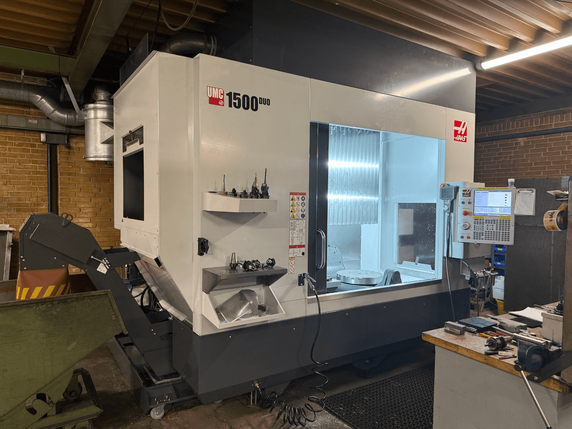 Haas UMC 1500DUO CNC staklės, vaizdas iš priekio, didelis langas, valdymo skydelis ir įrankių laikikliai ant lentynos.