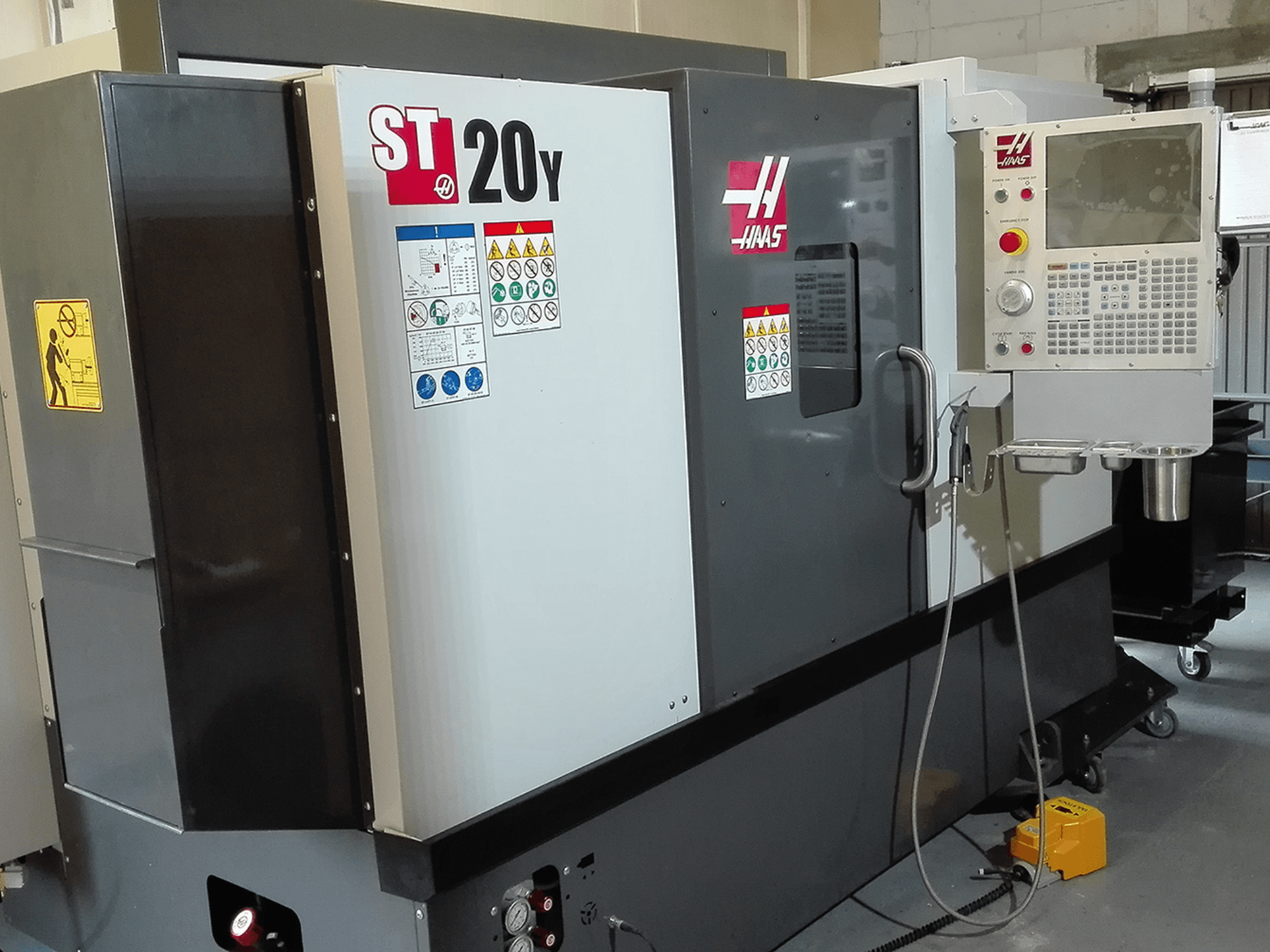 Haas ST20Y CNC tekinimo staklės iš priekio, su valdymo skydeliu, saugos etiketėmis ir aušinimo skysčio sistema.